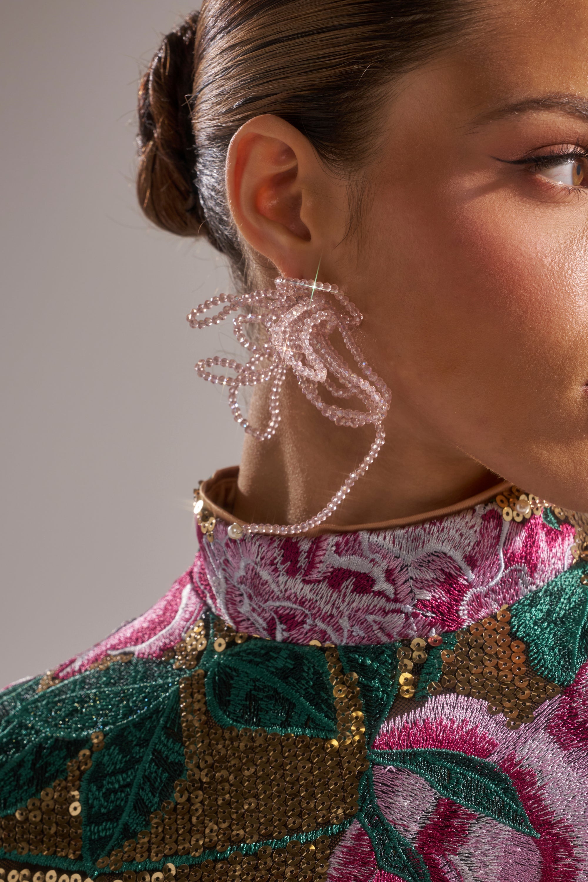 Close-up of a woman wearing the GIRL PWR EARRING—an abstract pink beaded statement piece. Her sleek bun and high-collared, sequin floral top frame the right side of her face. 5