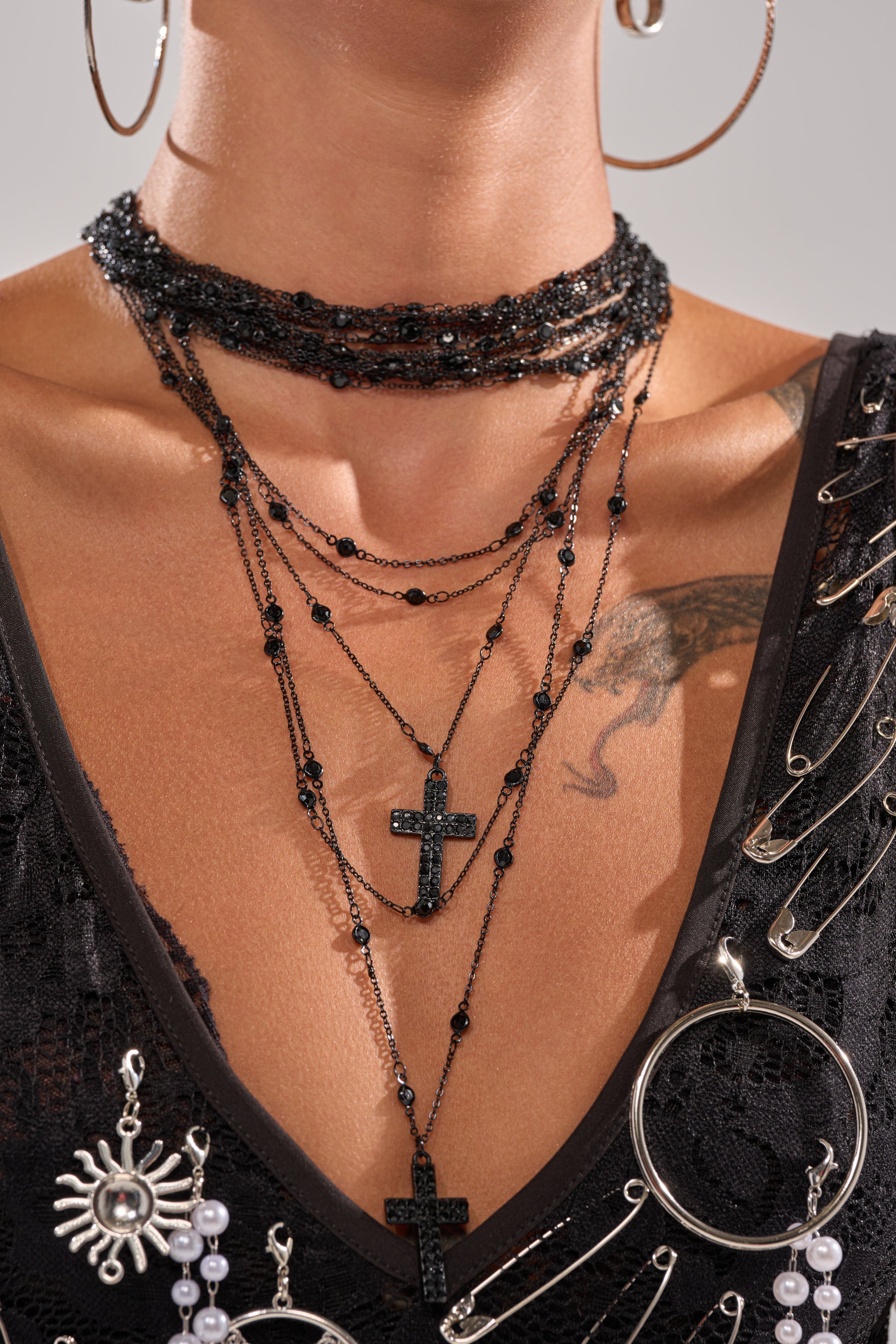 A person wears the DOLCE LAYERED NECKLACE with black cross pendants, large hoop earrings, and a black lace top detailed with safety pins and silver accents. Vampy jewelry adds edge, and a chest tattoo is visible. 1