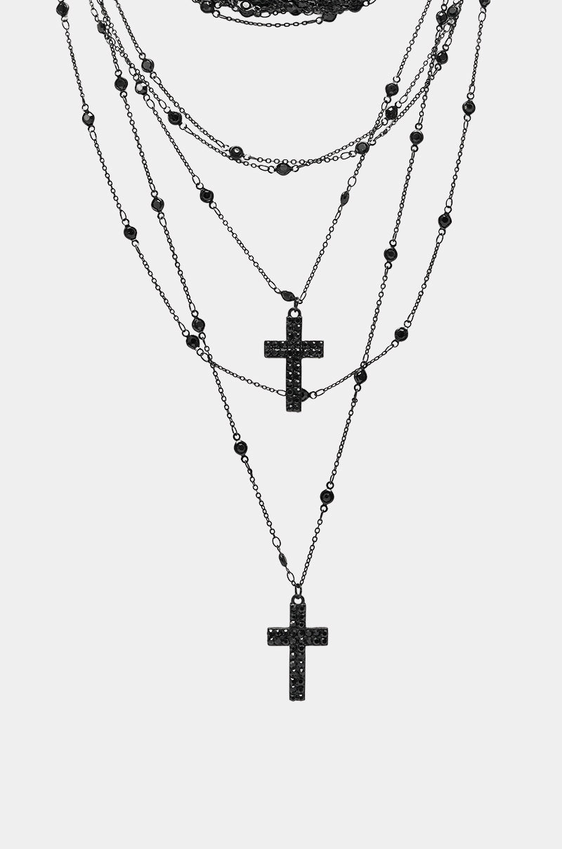 The DOLCE LAYERED NECKLACE features multiple black layered strands with black beaded accents and two gothic cross pendants adorned with small black stones, displayed against a plain white background. 3