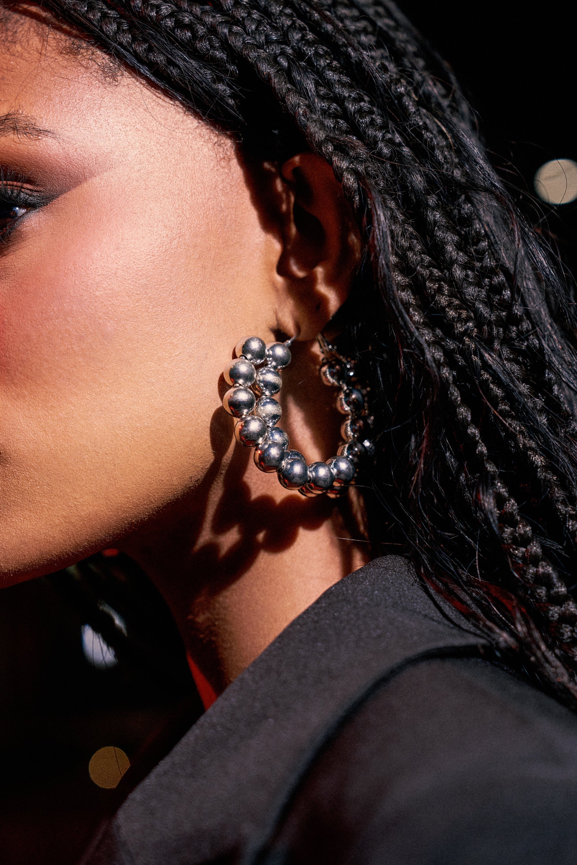 Close-up of a person with braided hair wearing the ROYALTY EARRING embellished with metallic beads. Smooth, glowing skin and a dark outfit pop against a softly blurred background.