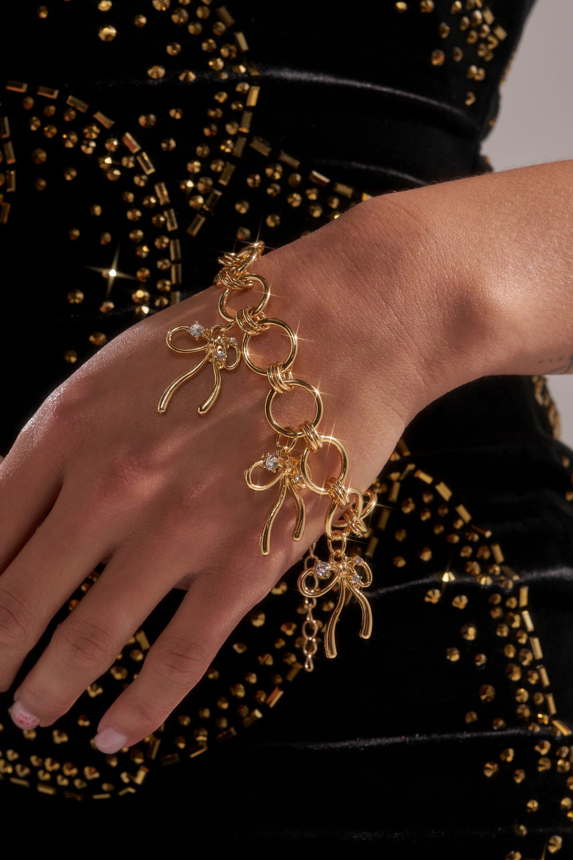 A close-up of a hand wearing the BOW TIME CHARM BRACELET—gold-tone with dangling charms and rhinestone accents—against a black velvet dress adorned with circular gold stud patterns. 1