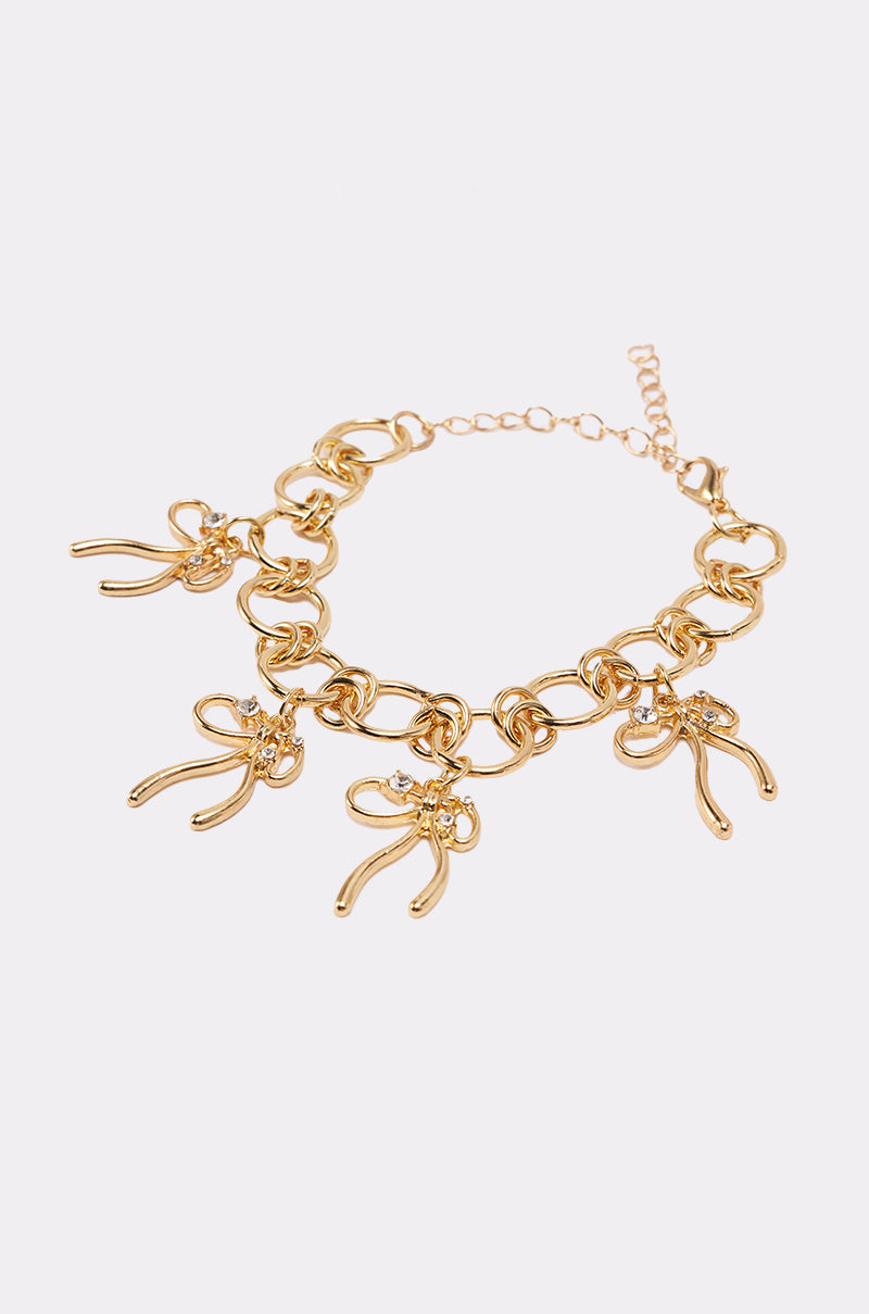 The BOW TIME CHARM BRACELET is a gold-tone bracelet with large links, featuring multiple bow charms, each detailed with a rhinestone at the center, shown against a plain white background. 2