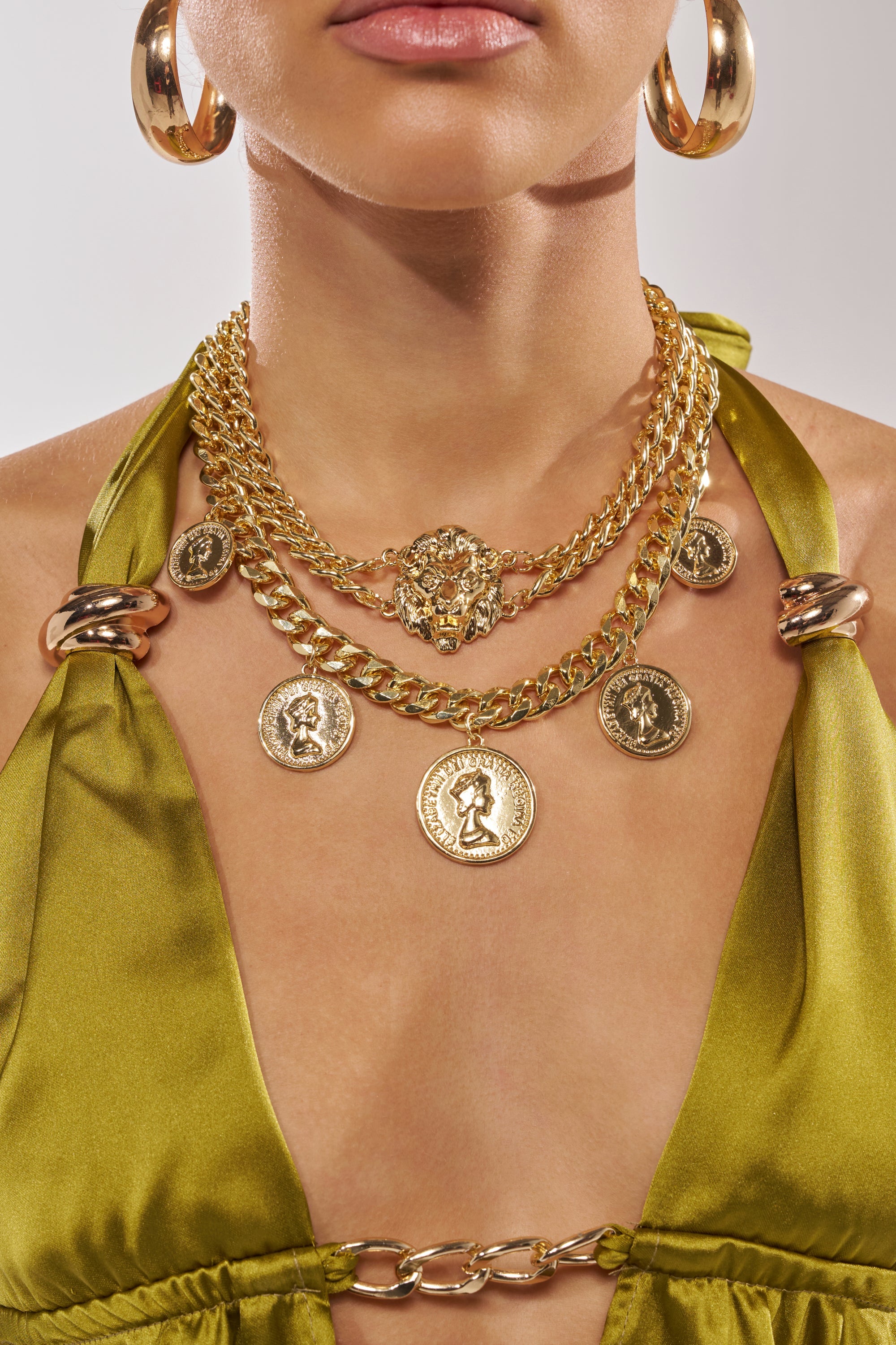 A woman wears large gold hoop earrings and the AM WHO I THINK I AM NECKLACE—a chunky layered coin necklace with pendants—paired with a shiny green halter top featuring chain details at the neckline. 1