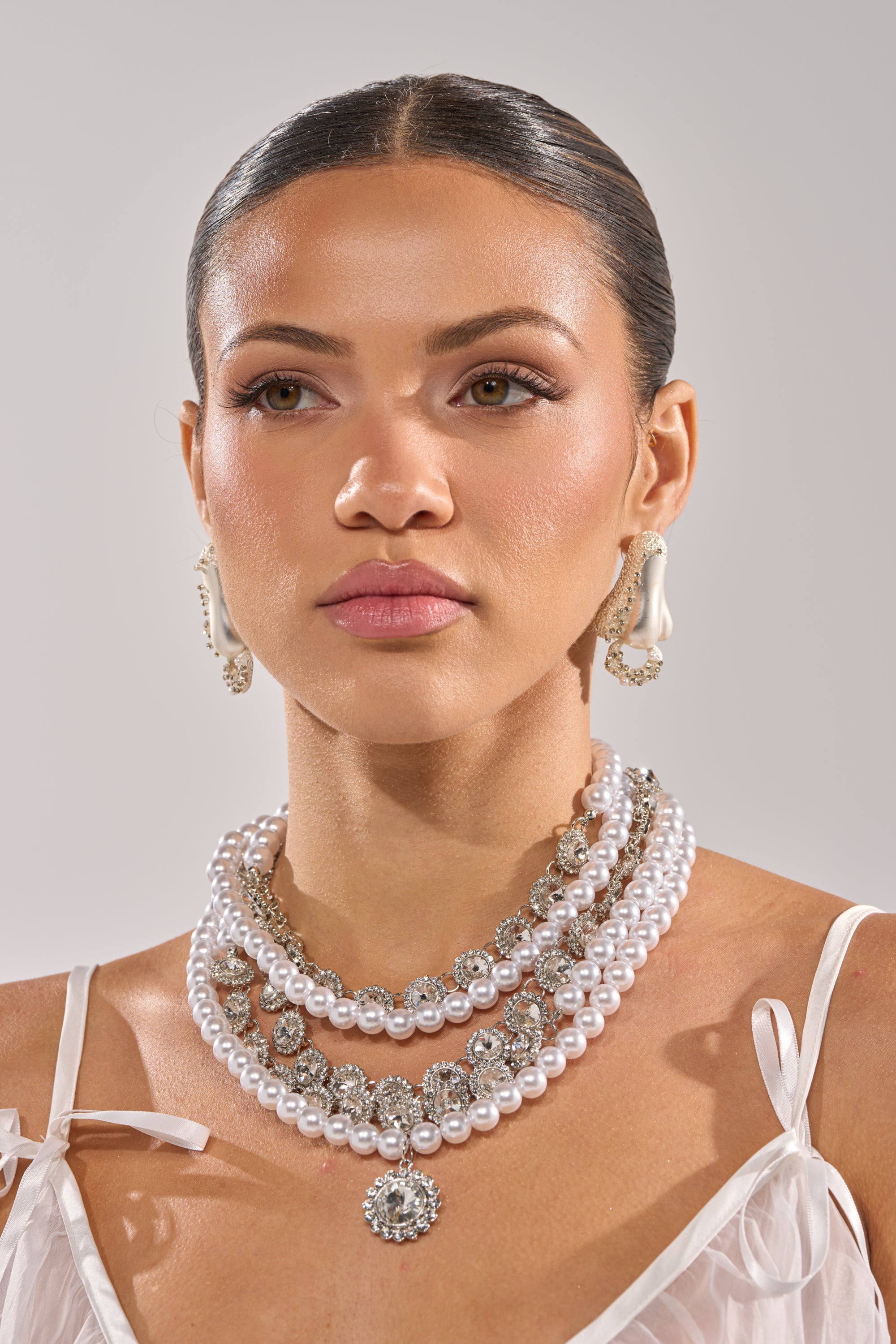 A woman with slicked-back hair wears pearl earrings and the AFFORD IT NECKLACE featuring jeweled accents, styled with a white spaghetti-strap top against a plain, light background. 4