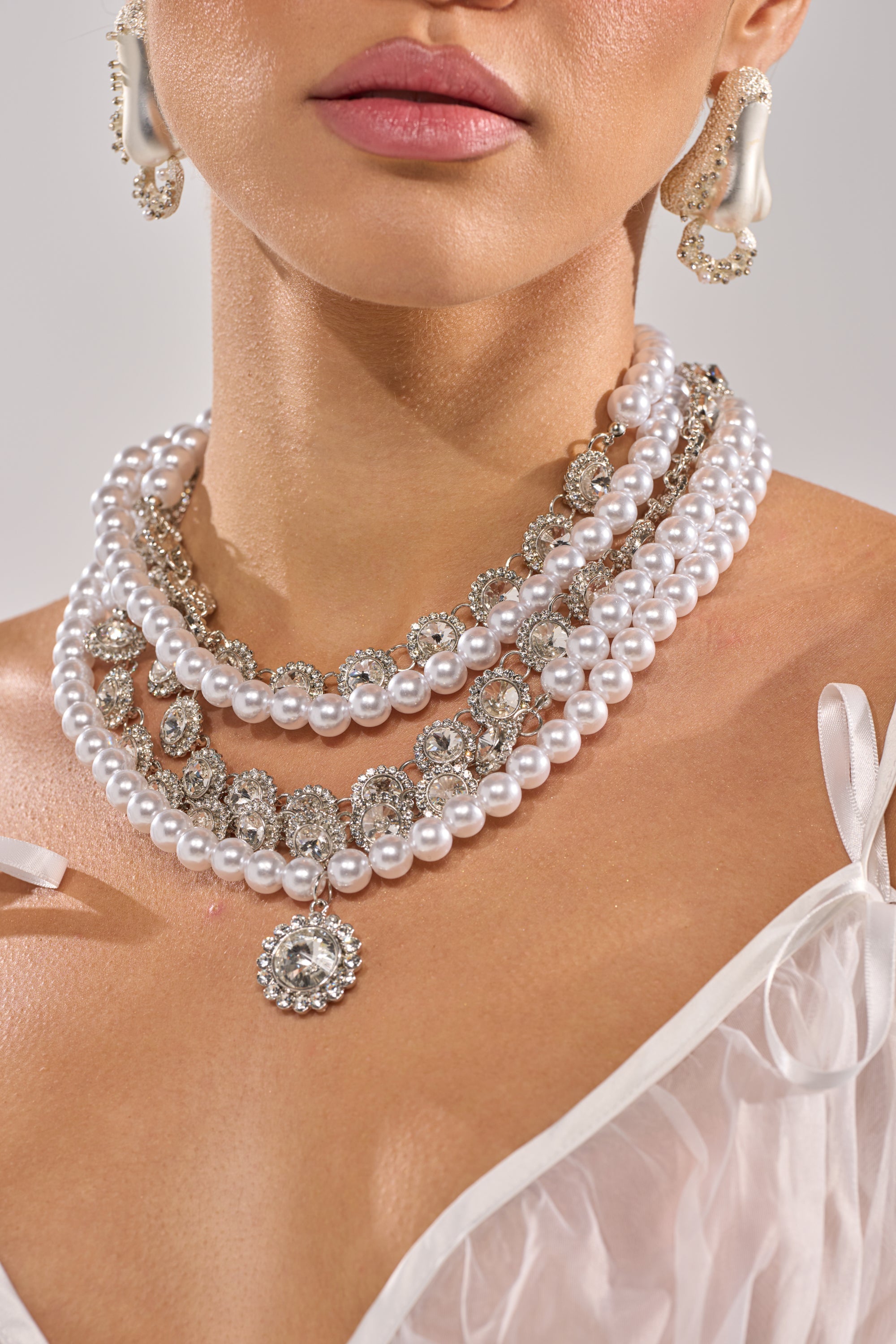 A woman wears the AFFORD IT NECKLACE, a vintage-inspired layered piece with pearls and rhinestones, styled with a jeweled pendant and ornate earrings. She’s dressed in a sheer white top with delicate straps, showing her neck and shoulders. 1