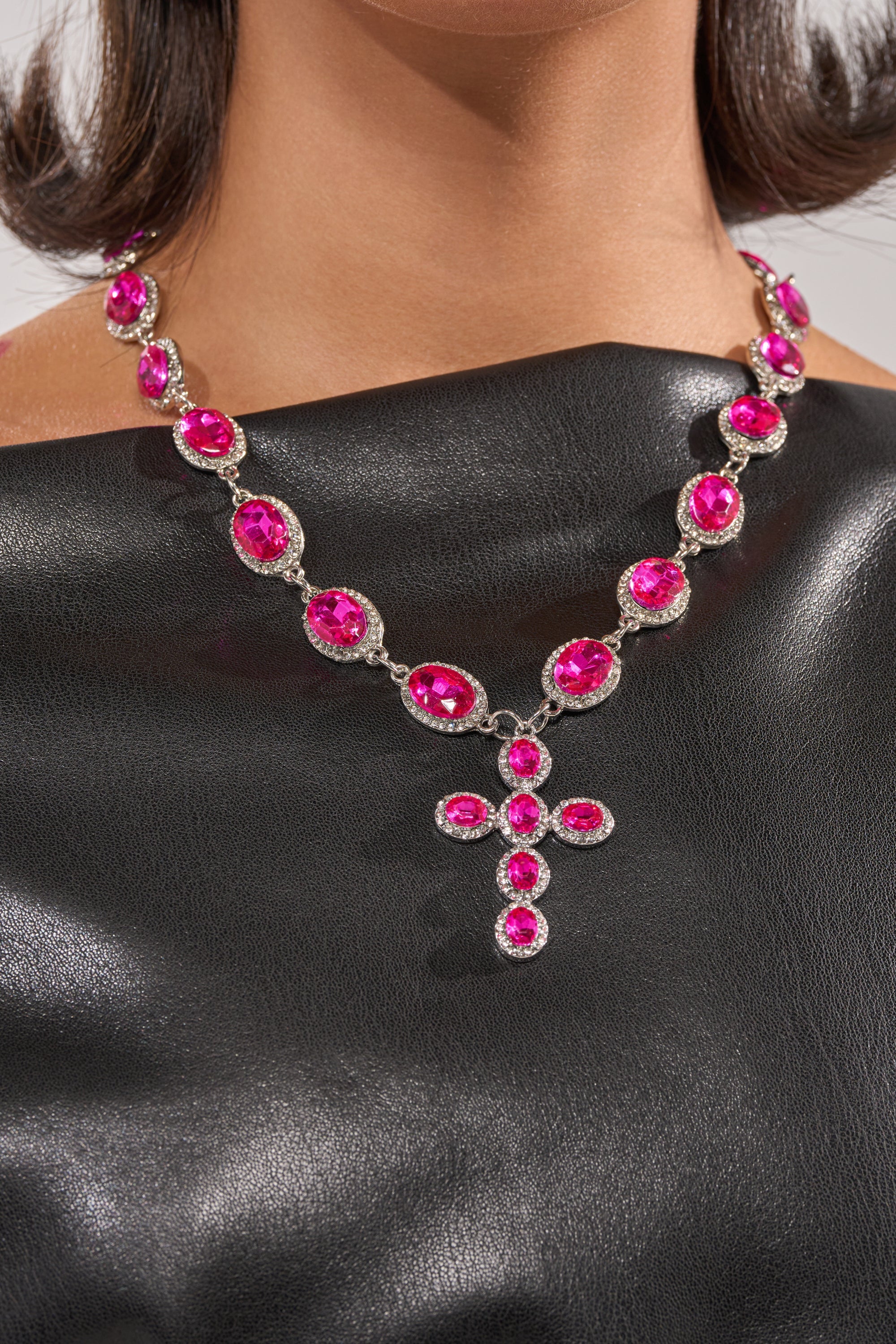 A woman in a shiny black off-the-shoulder top wears the NELLY NECKLACE, a hot pink rhinestone necklace with a cross pendant and matching stones on a delicate crystal station chain. 2