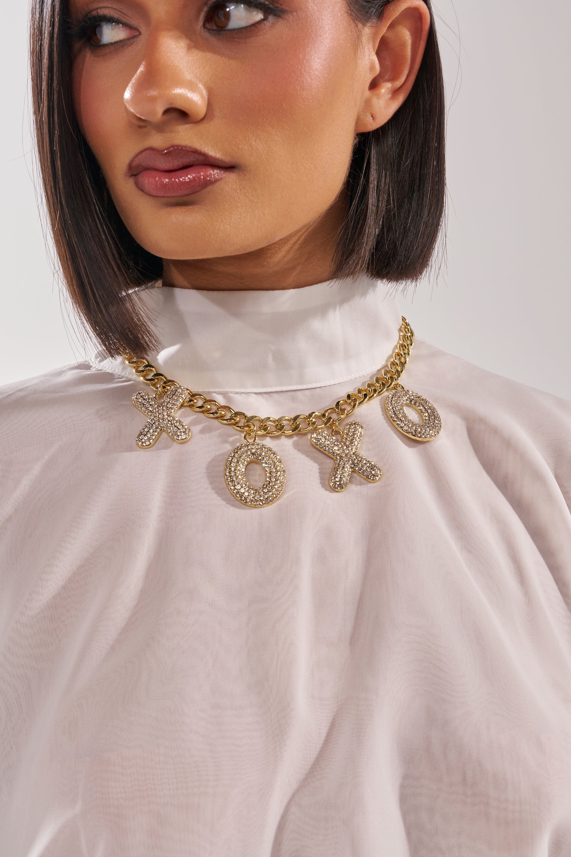 A woman wears a sheer white high-neck blouse and the KISSES NECKLACE, a chunky gold pavé curb chain adorned with large crystal XO charms. Only her face and upper torso are visible. 2
