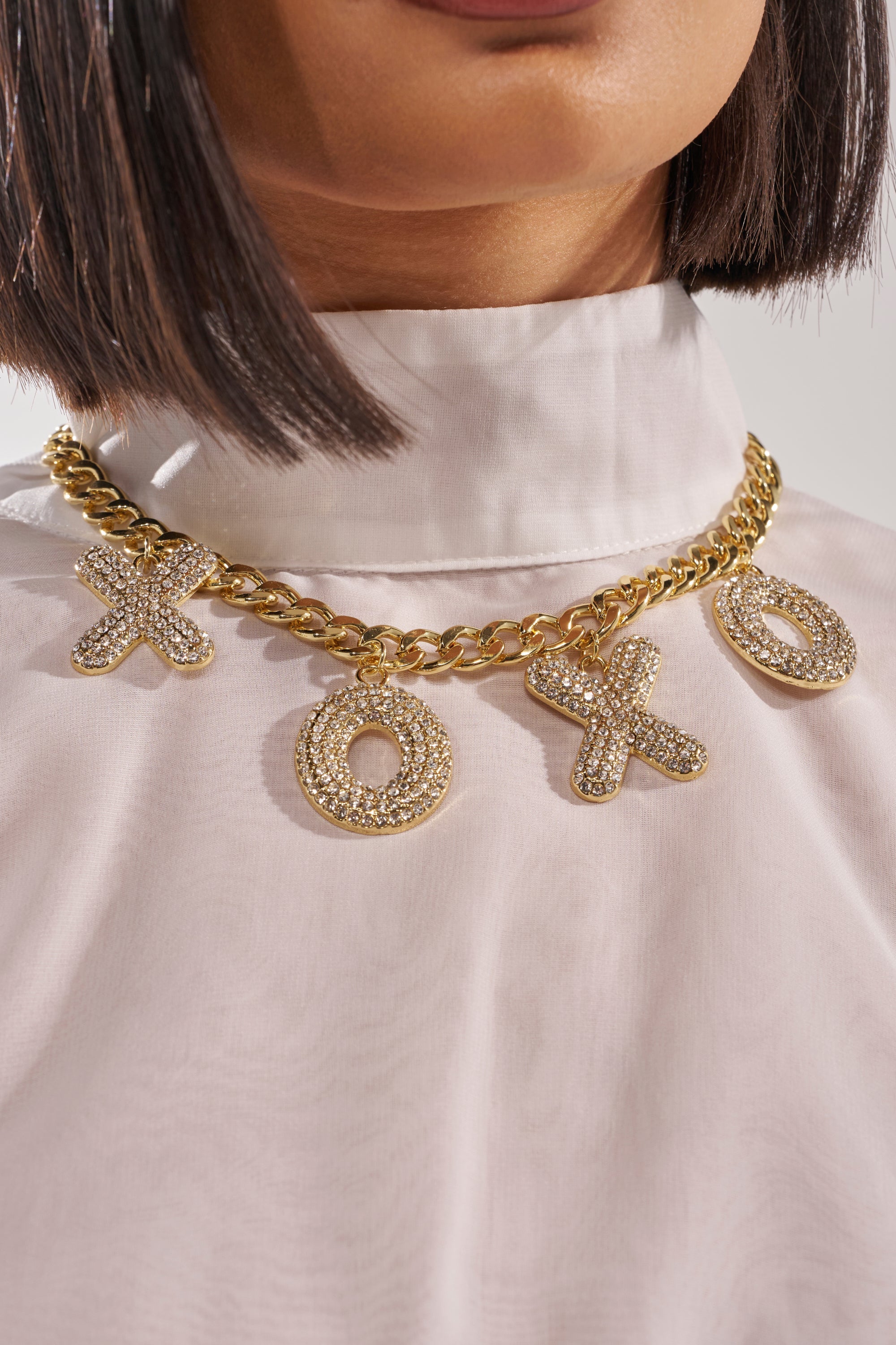 A woman wears a white high-neck blouse paired with the KISSES NECKLACE, featuring bold crystal XO letter charms. 3