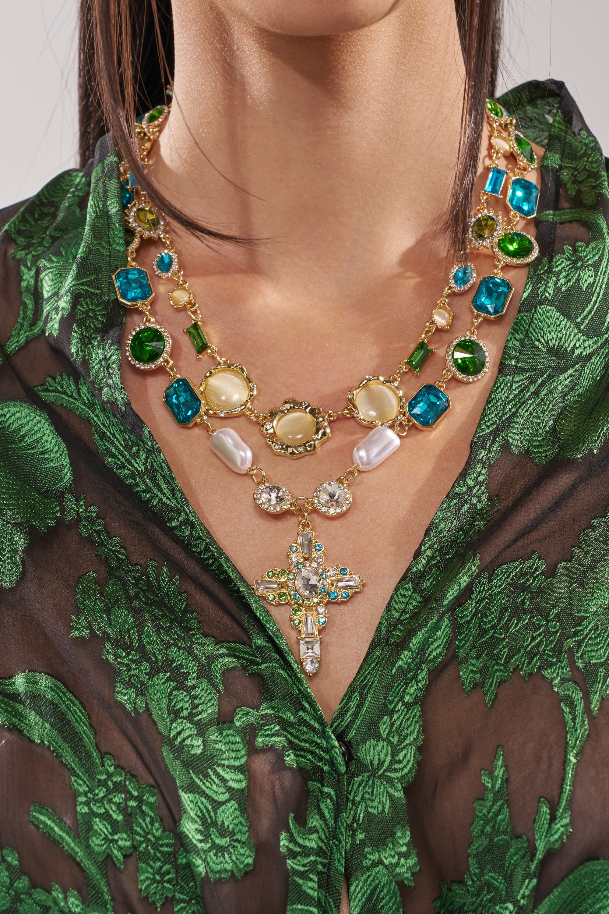 A woman wears a sheer black blouse with green floral patterns, accessorized with the ON A YACHT NECKLACE—a vintage-inspired jeweled cross pendant adorned with colorful gemstones, pearls, and a statement crystal cross. 1