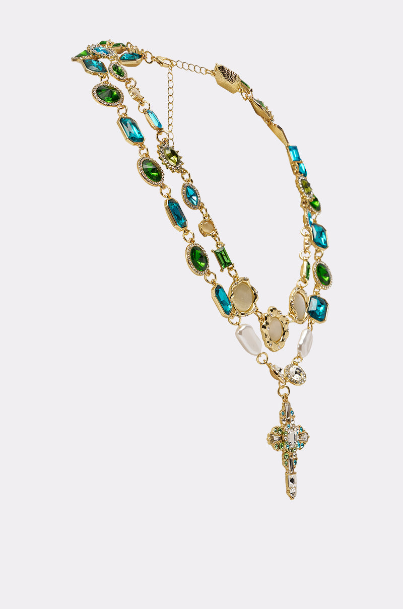 The ON A YACHT NECKLACE is a gold vintage-inspired piece with blue, green, and white gemstones, layered with an ornate jeweled cross pendant, shown against a simple light background. 2