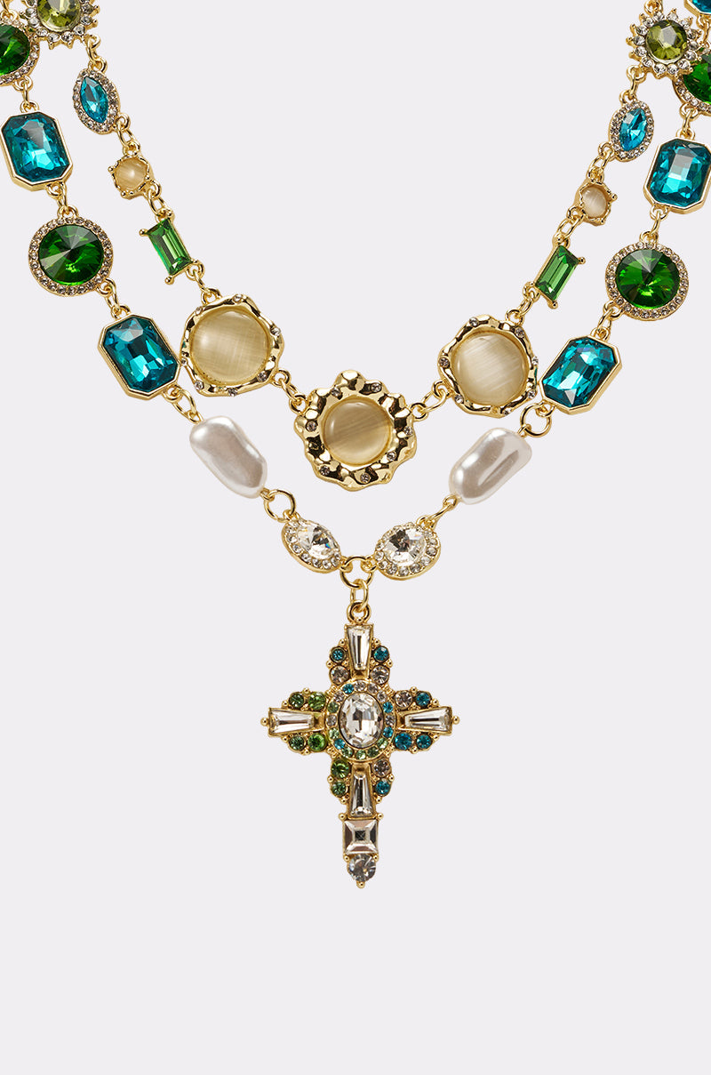 The ON A YACHT NECKLACE features a vintage-inspired design with green and blue gemstones, pearl-like beads, circular gold accents, and a central crystal statement cross. 3