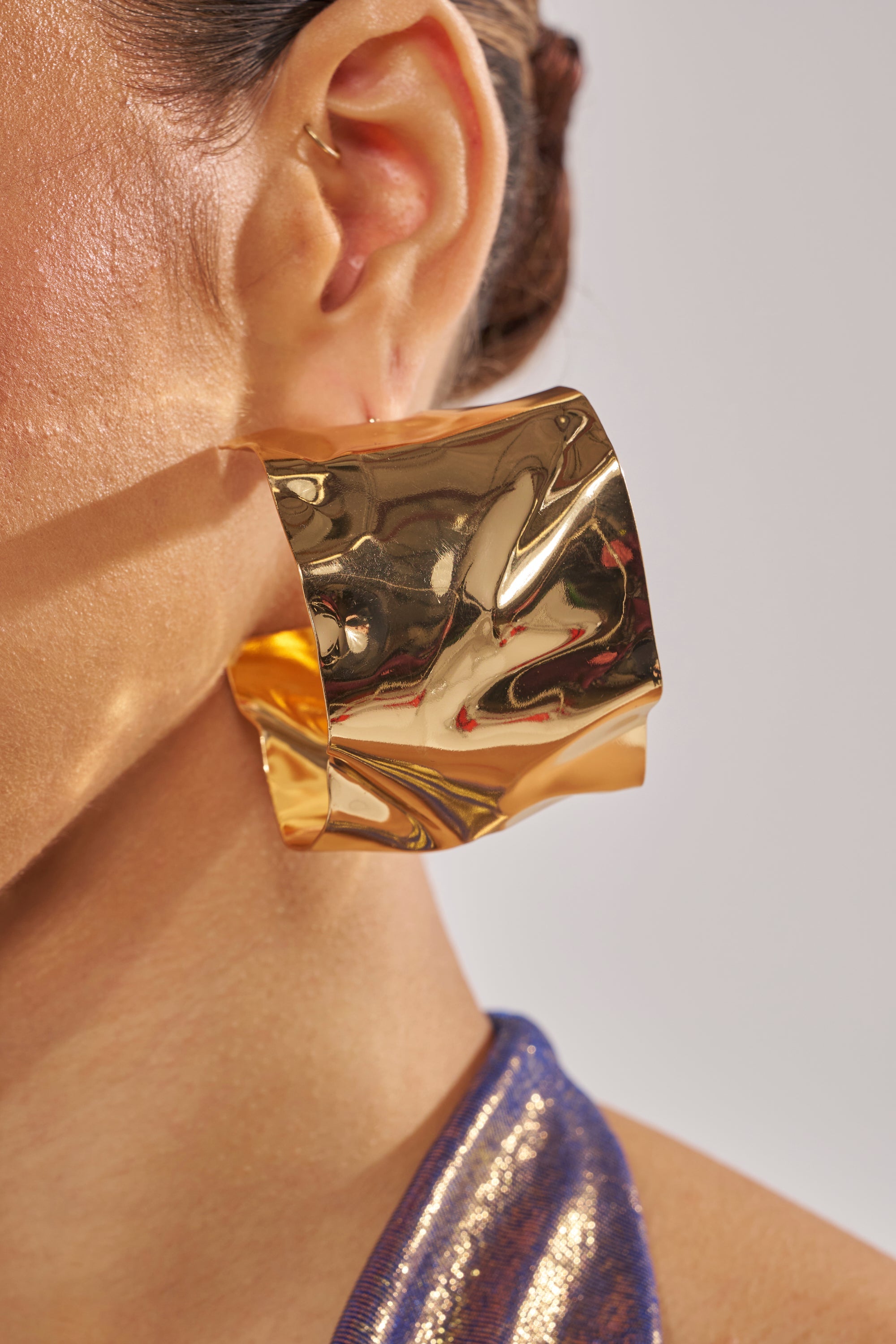 Close-up of a person's ear wearing the BENT OUTTA SHAPE EARRING—oversized hoops with a sculptural, crinkled gold finish. The person has tied-back hair and wears a shiny metallic blue top. 4