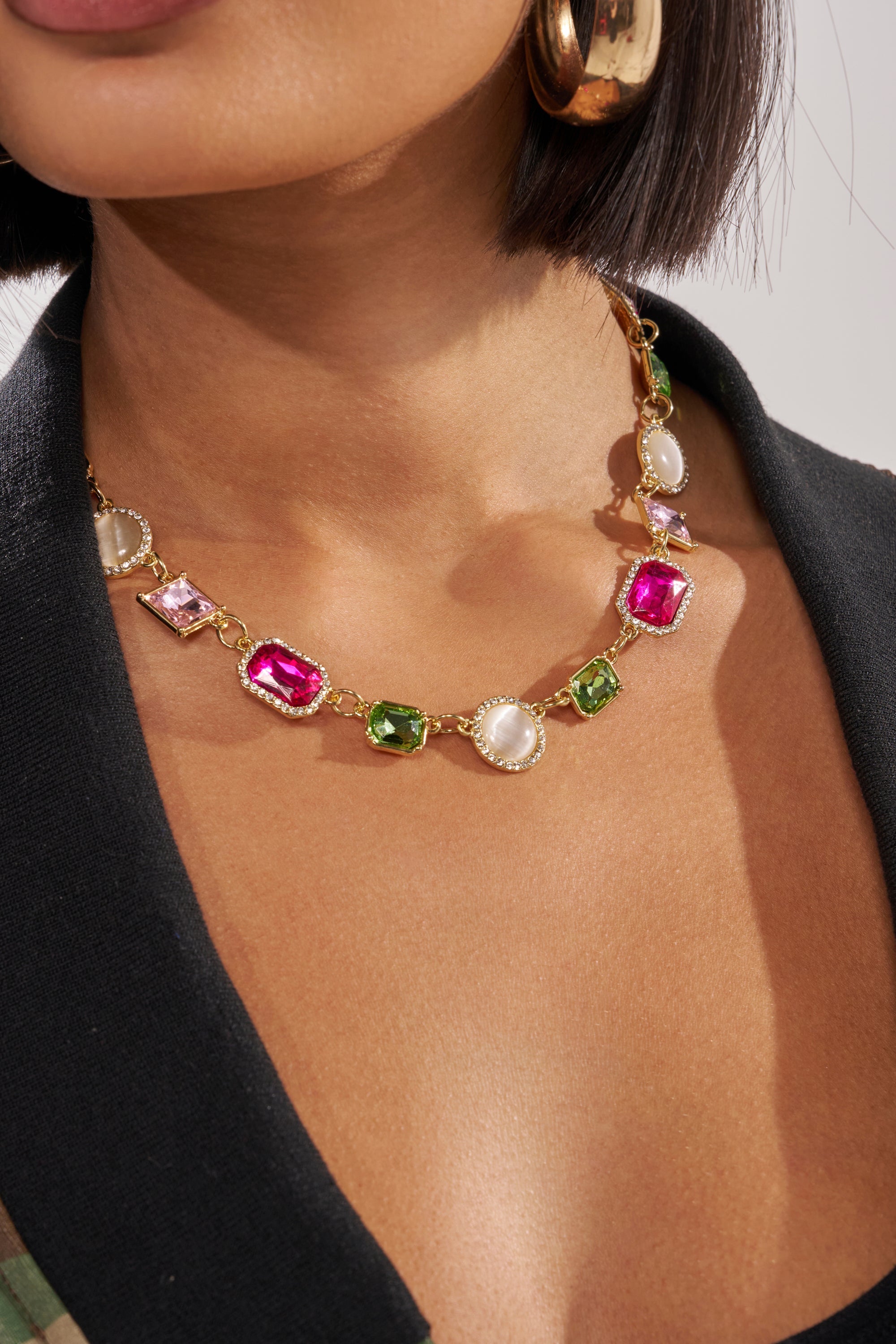 A close-up of a woman wearing the WHO DO IT LIKE ME NECKLACE, a multicolor gem piece with gold hardware and pavé accents—pink, green, and pearl-like stones—paired with a black blazer and large gold hoop earring. 1