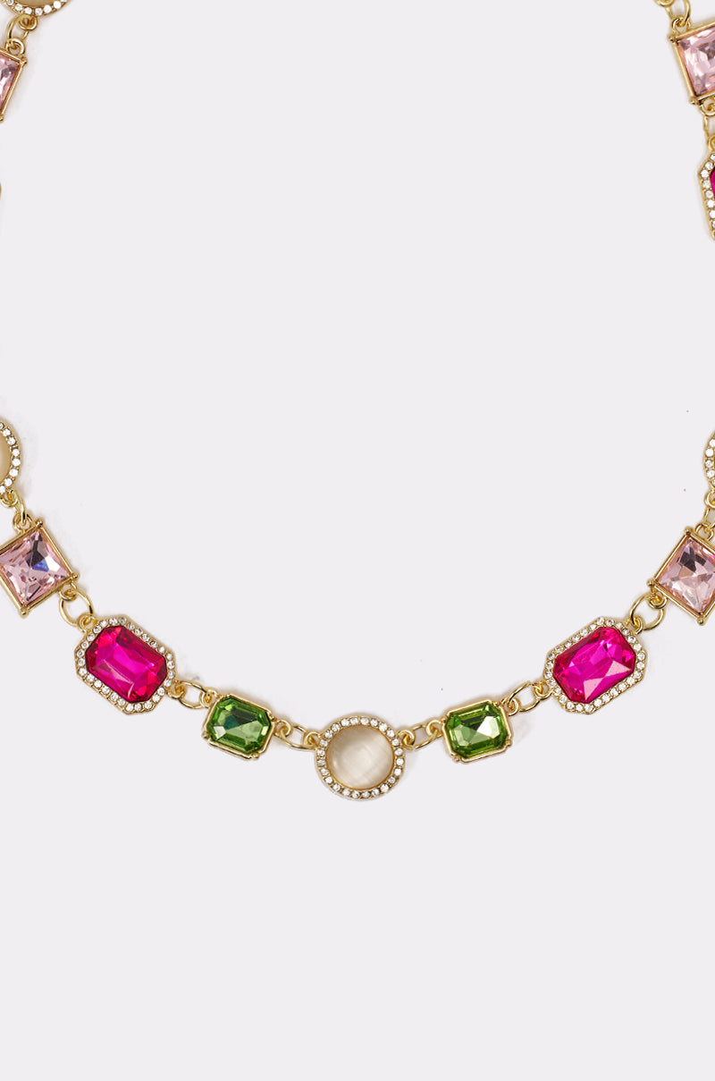 The WHO DO IT LIKE ME NECKLACE features rectangular pink, green, and clear gemstones with a central oval white stone in decorative bezels, accented by elegant gold hardware, all displayed against a white background. 2