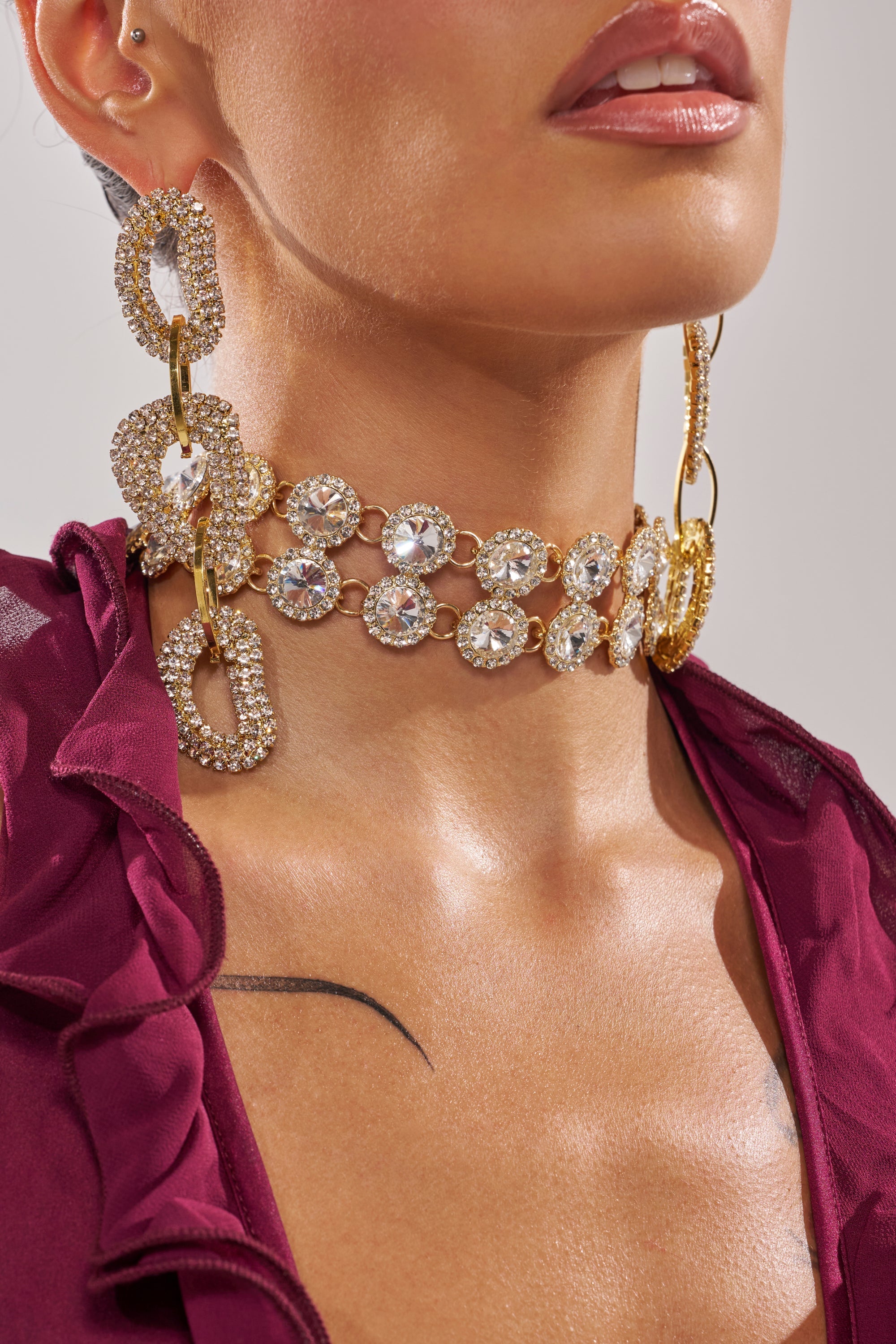 A close-up of a person wearing the RIRI NECKLACE with matching earrings, styled with a deep burgundy ruffled top and a visible collarbone tattoo against a neutral background. 1