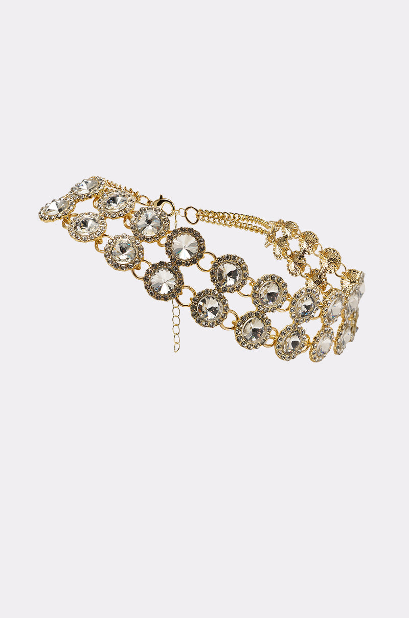 The RIRI NECKLACE is a gold-tone crystal statement choker with three rows of large, round, clear gemstones in ornate gold settings for an elegant, luxurious look. 2