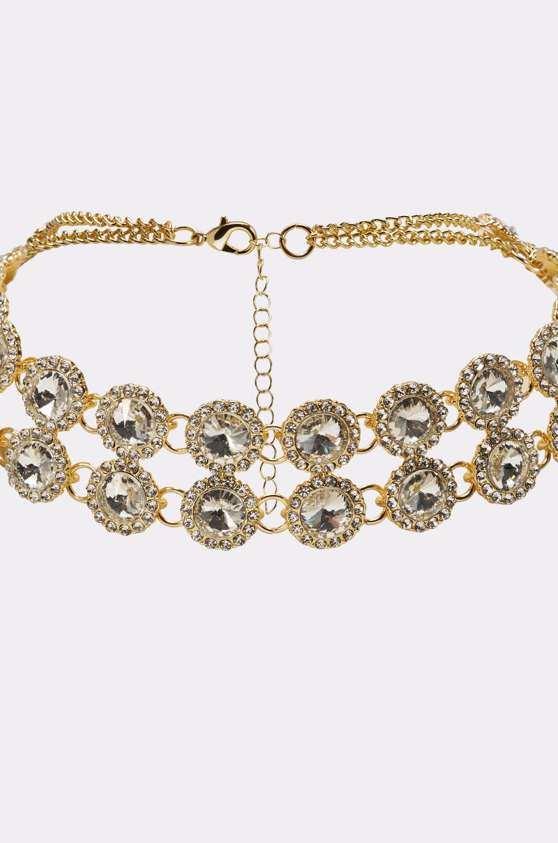 The RIRI NECKLACE is a gold-tone statement choker with two rows of clear, circular gemstones bordered by smaller crystals and an adjustable clasp chain at the back. 3