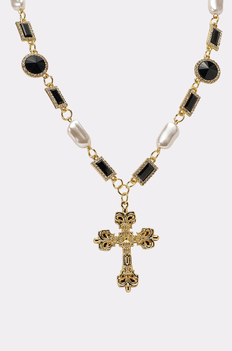 The WITH THE TIMES NECKLACE is a gold-tone cross necklace with an ornate pendant, featuring gothic-glam black gemstones and white pearls set along the chain, shown on a plain light background. 4