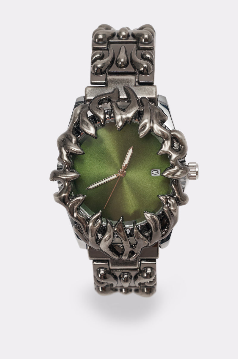 The IN MY VIBE WATCH features a silver sculptural link band with claw-inspired details, a spiked bezel, a bold green dial, silver hands, and a date window. 1