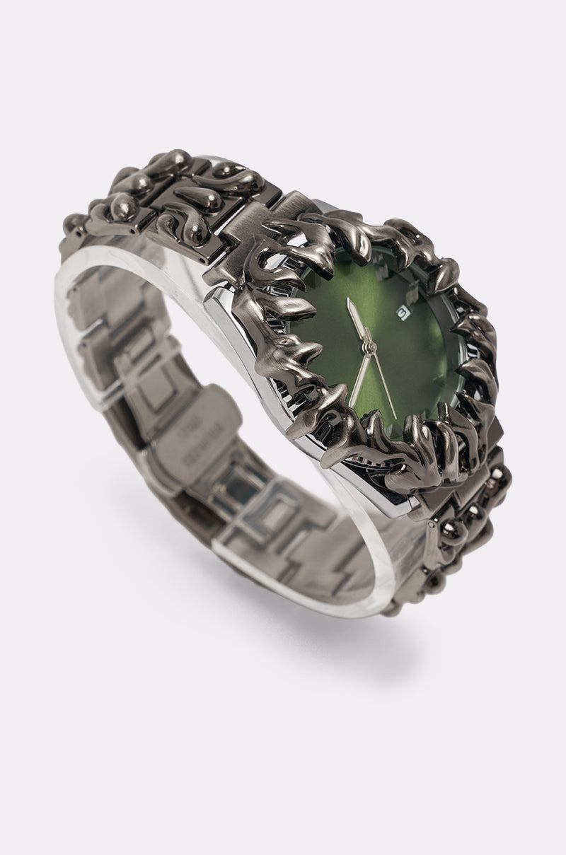 The IN MY VIBE WATCH features a silver sculptural link band and a spiked, flame-like bezel framing its bold green dial, finished with sleek silver hour and minute hands. 2