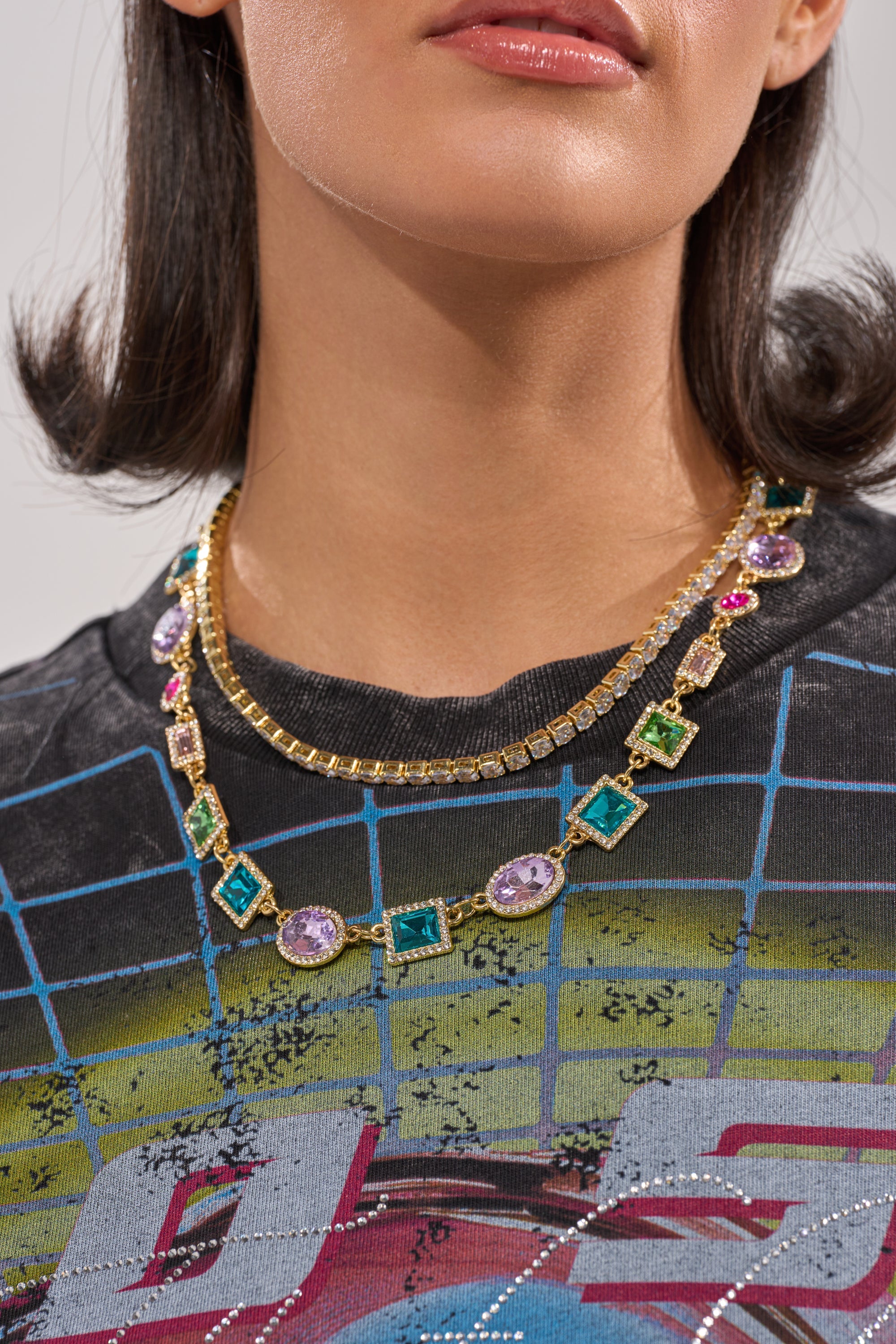 A woman wears a black graphic T-shirt with a grid pattern and the RAY NECKLACE—a statement piece adorned with colorful gemstones and small clear stones. Only her lips, chin, and neckline are visible. 1