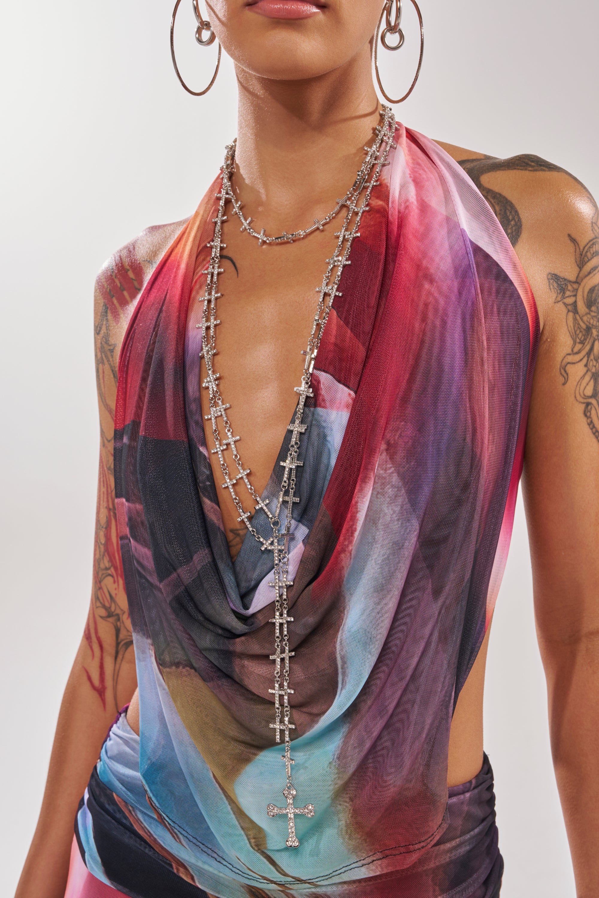 A person wearing a colorful draped top, large hoop earrings, visible arm tattoos, and the TELL ME BOUT IT NECKLACE layered with other chains. The image captures them from shoulders to waist. 1