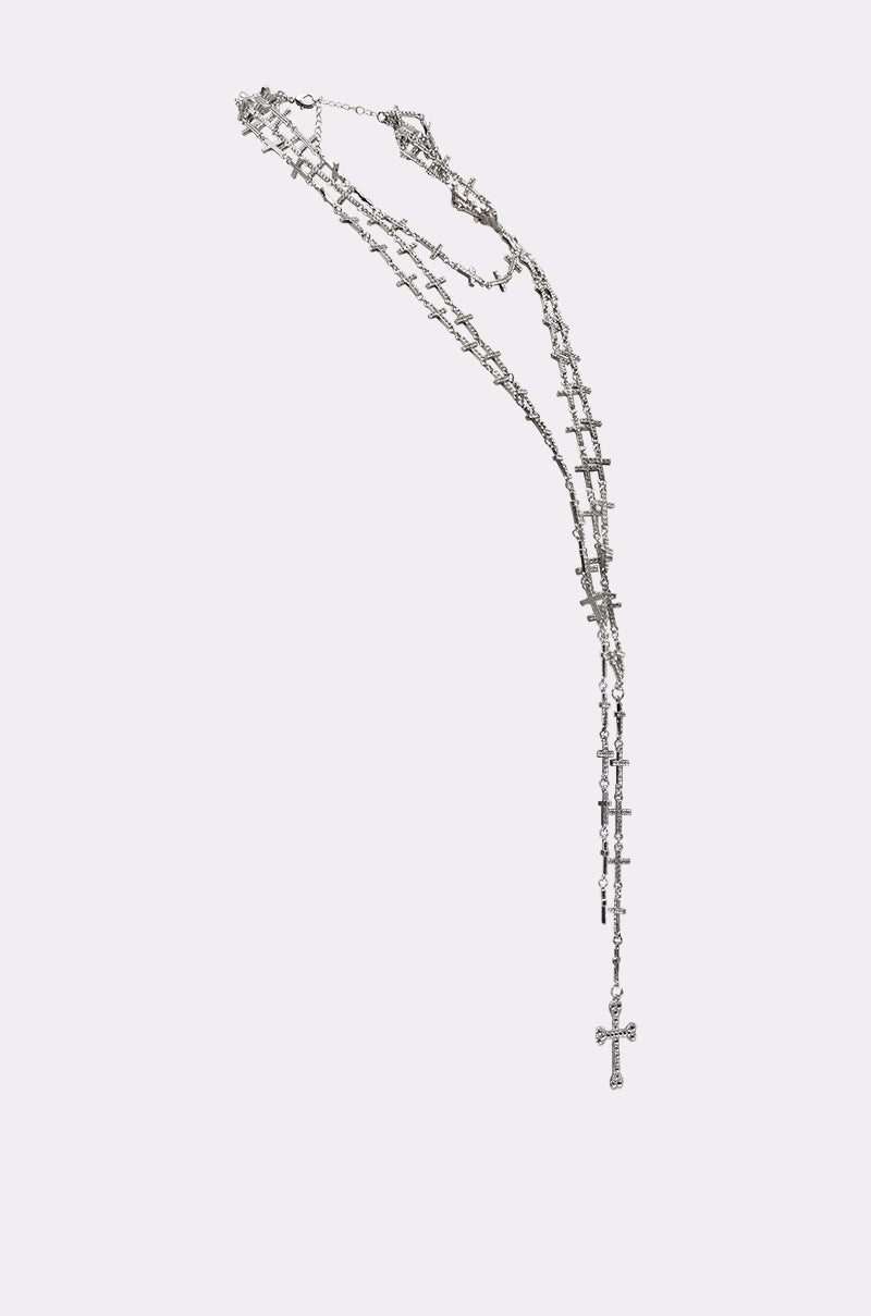 The TELL ME BOUT IT NECKLACE features layered silver chains with small cross pendants and a larger statement cross at the end, displayed against a plain light background. 2