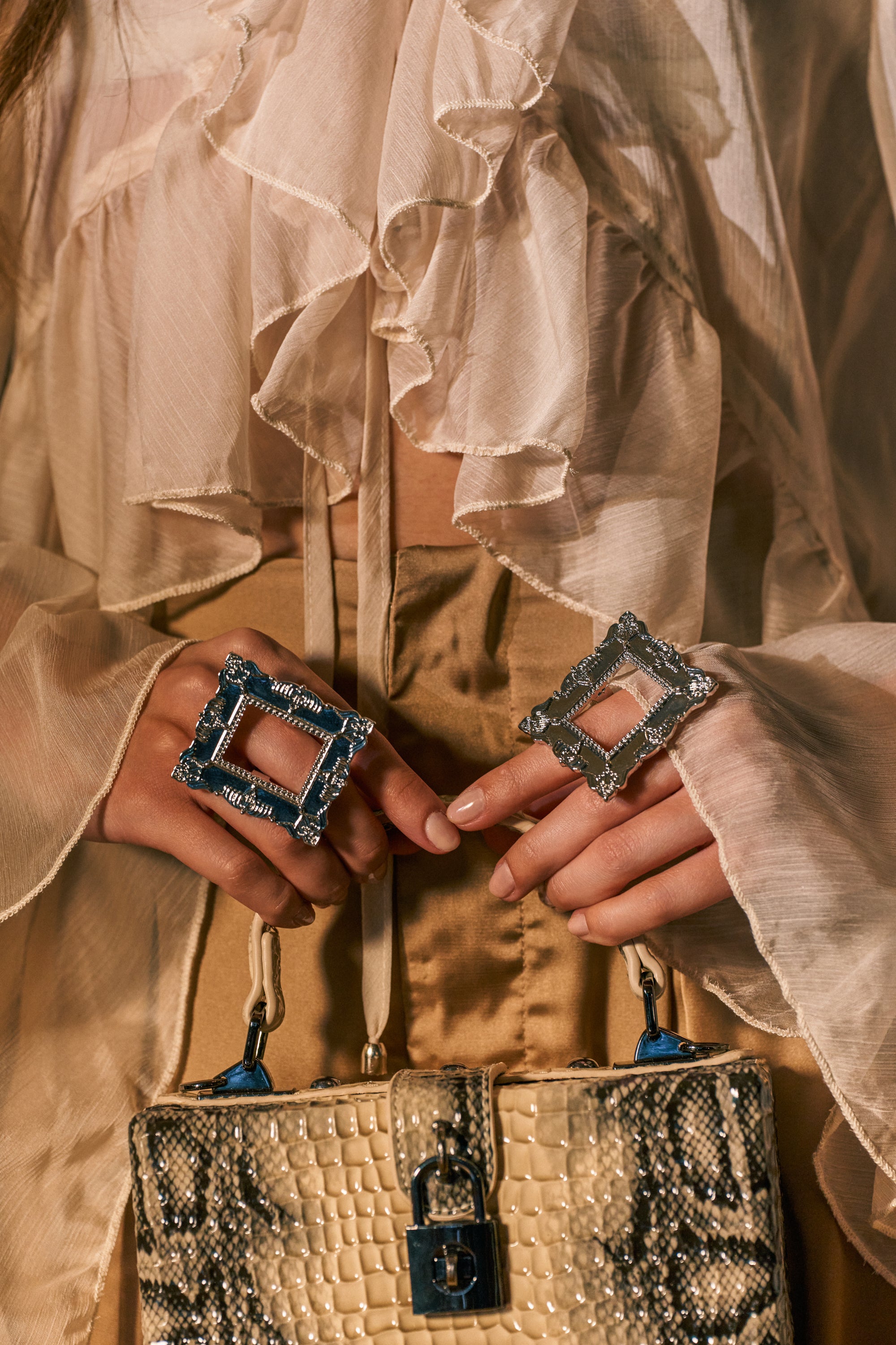 A person wearing a ruffled, sheer blouse and satin pants holds a textured handbag. Adorning their fingers are two large, square, ornate statement rings from the FRAMED MIND COCKTAIL RING SET, including a striking silver picture frame design. The overall style appears luxurious and fashionable.