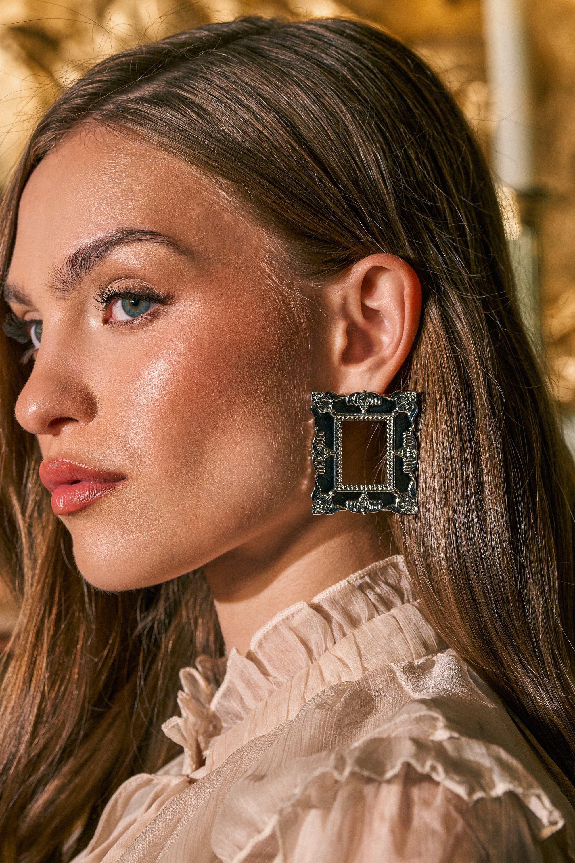 A woman with long brown hair wears a ruffled cream blouse and the FRAMED MIND EARRING, a large, square, baroque-inspired statement piece. She gazes to the side with a soft, neutral expression against a warmly lit, out-of-focus background.
