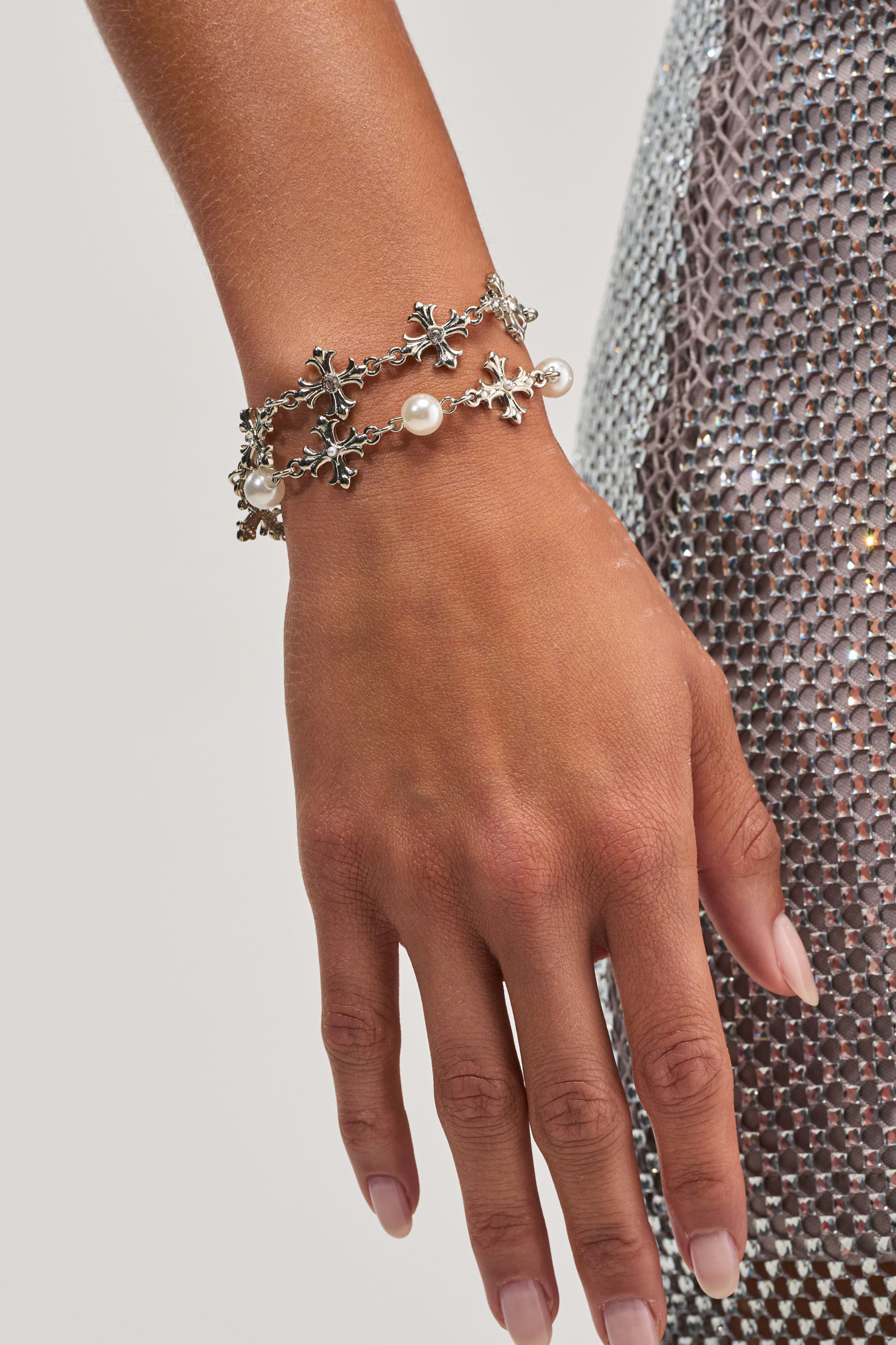A close-up of a hand wearing the TOO LIT BRACELET SET with silver-tone pearls and floral details, set against a silver sequined dress. The nails have a natural, glossy finish.