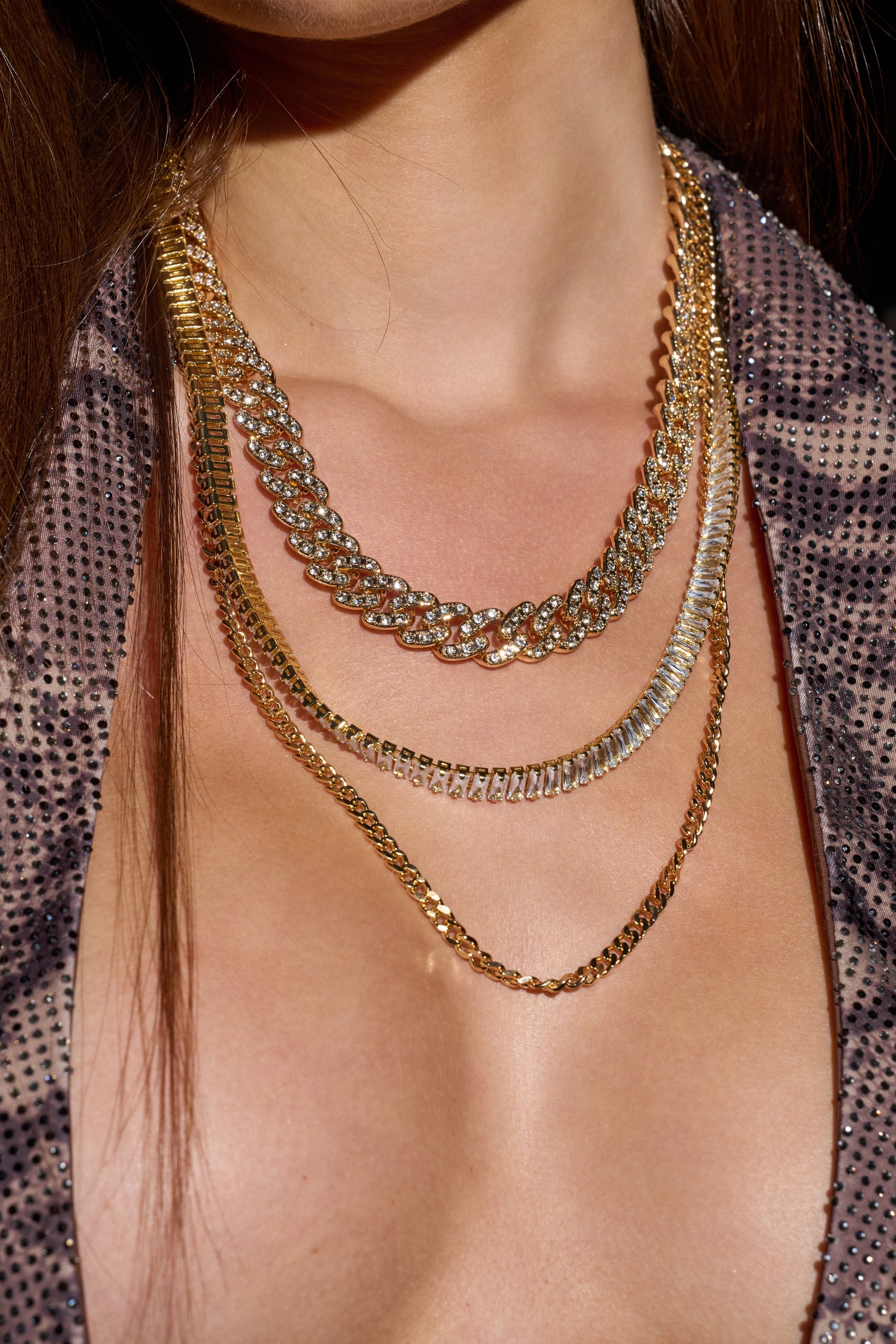 A close-up of a woman wearing a sparkly, deep-neck top showcases the ICY LIFE NECKLACE—three layered gold chains, including one with rhinestones—elegantly draped against her bare skin.