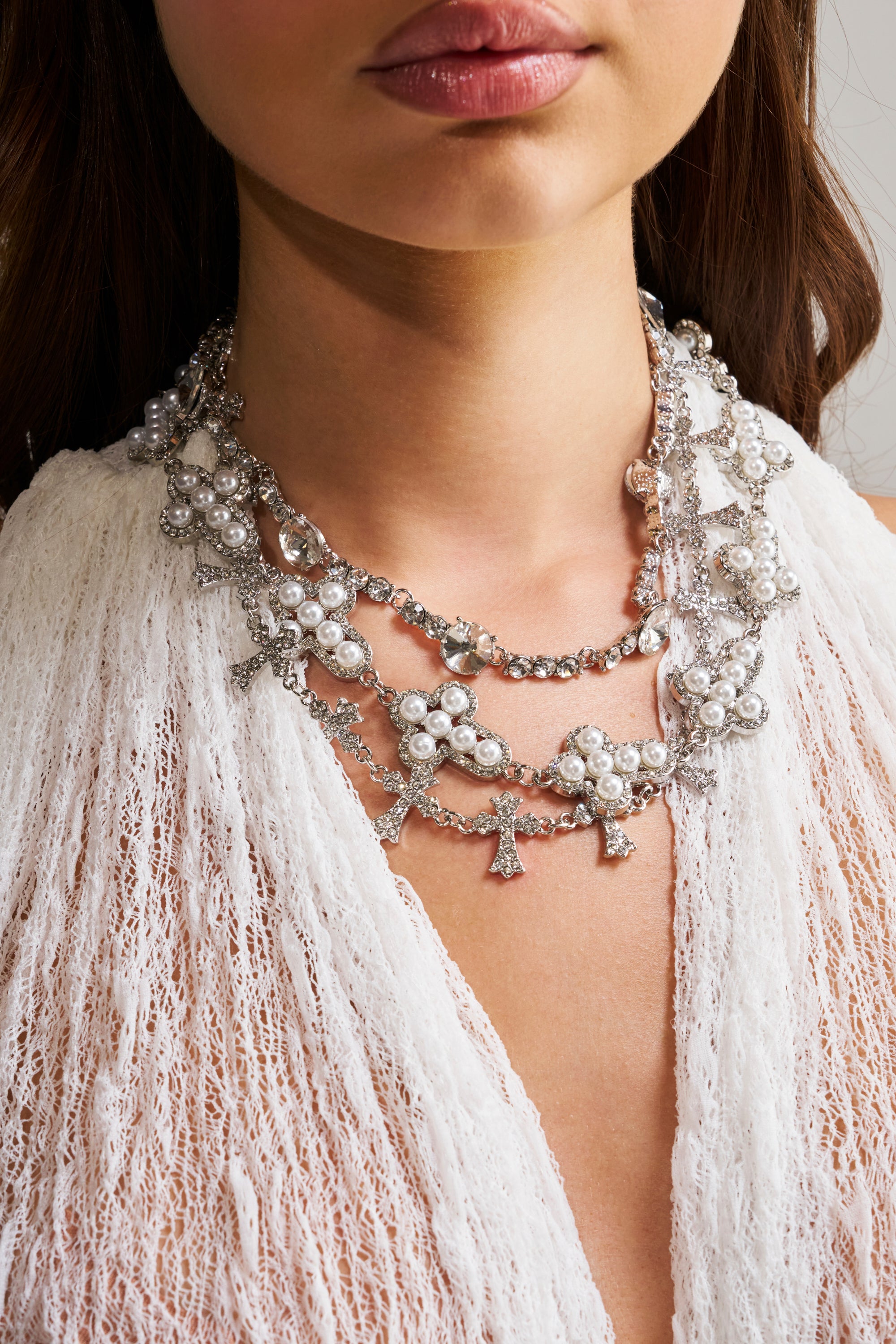 A woman wearing a textured white top showcases the IM OUTSIDE NECKLACE SET, a layered statement necklace adorned with pearls and rhinestone cross charms for extra sparkle.