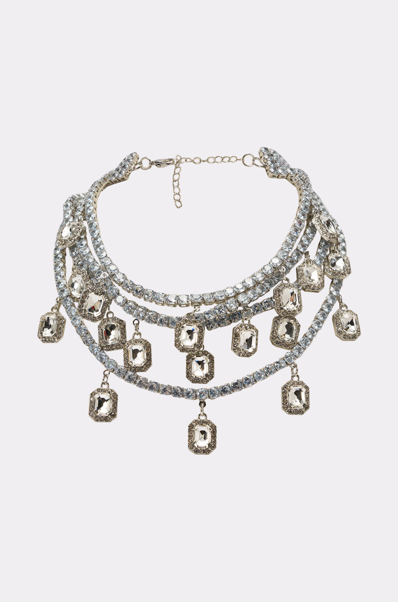 The BELLE LAYERED NECKLACE is a multi-layered silver statement piece with sparkling stones and nine dangling emerald-cut crystal pendants, shown on a white background.