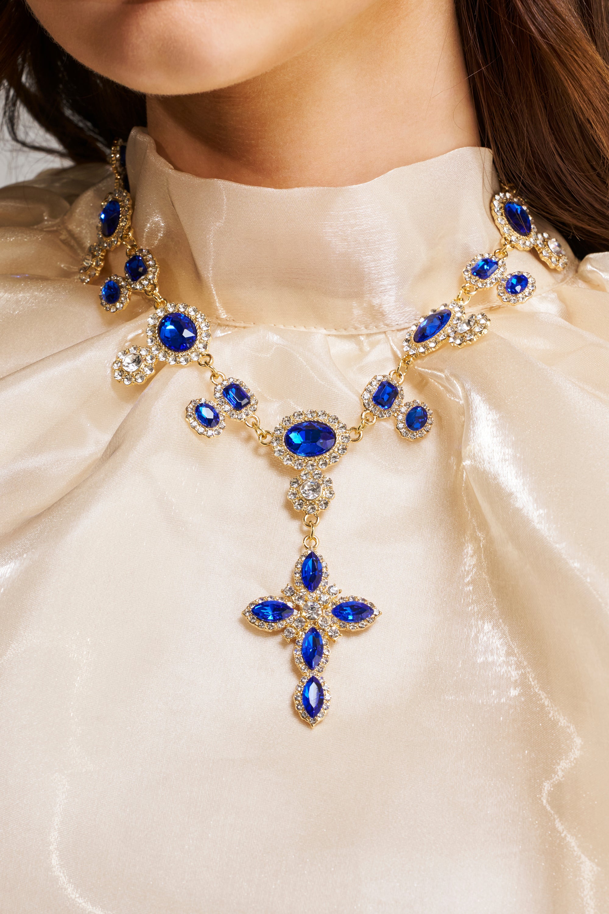 A woman wears the U DONT GET IT NECKLACE, an ornate gold statement necklace with large blue gemstones, clear crystals, and a striking sapphire cross pendant, paired with a shiny cream high-neck blouse.