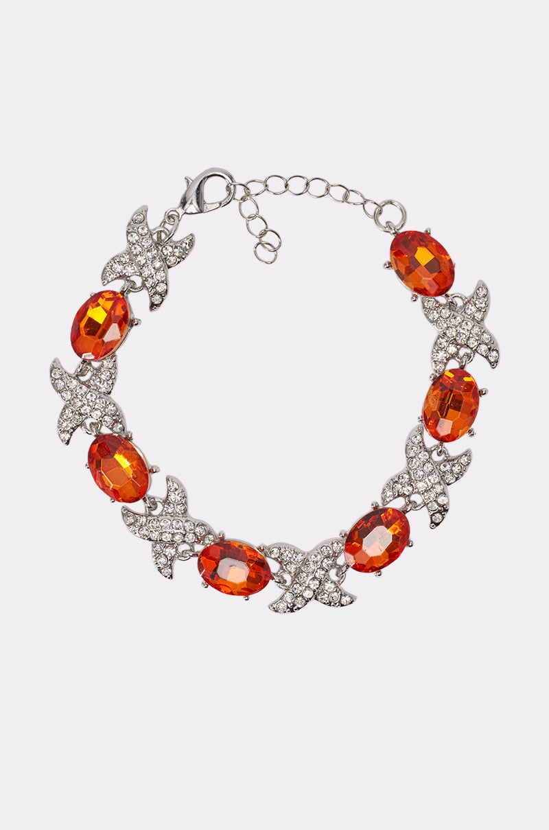 The ORANGE YA GLAD LINK BRACELET is a silver bracelet with alternating oval orange gemstones and X-shaped links adorned with clear rhinestones, displayed on a plain white background—an elegant accessory for summer parties.