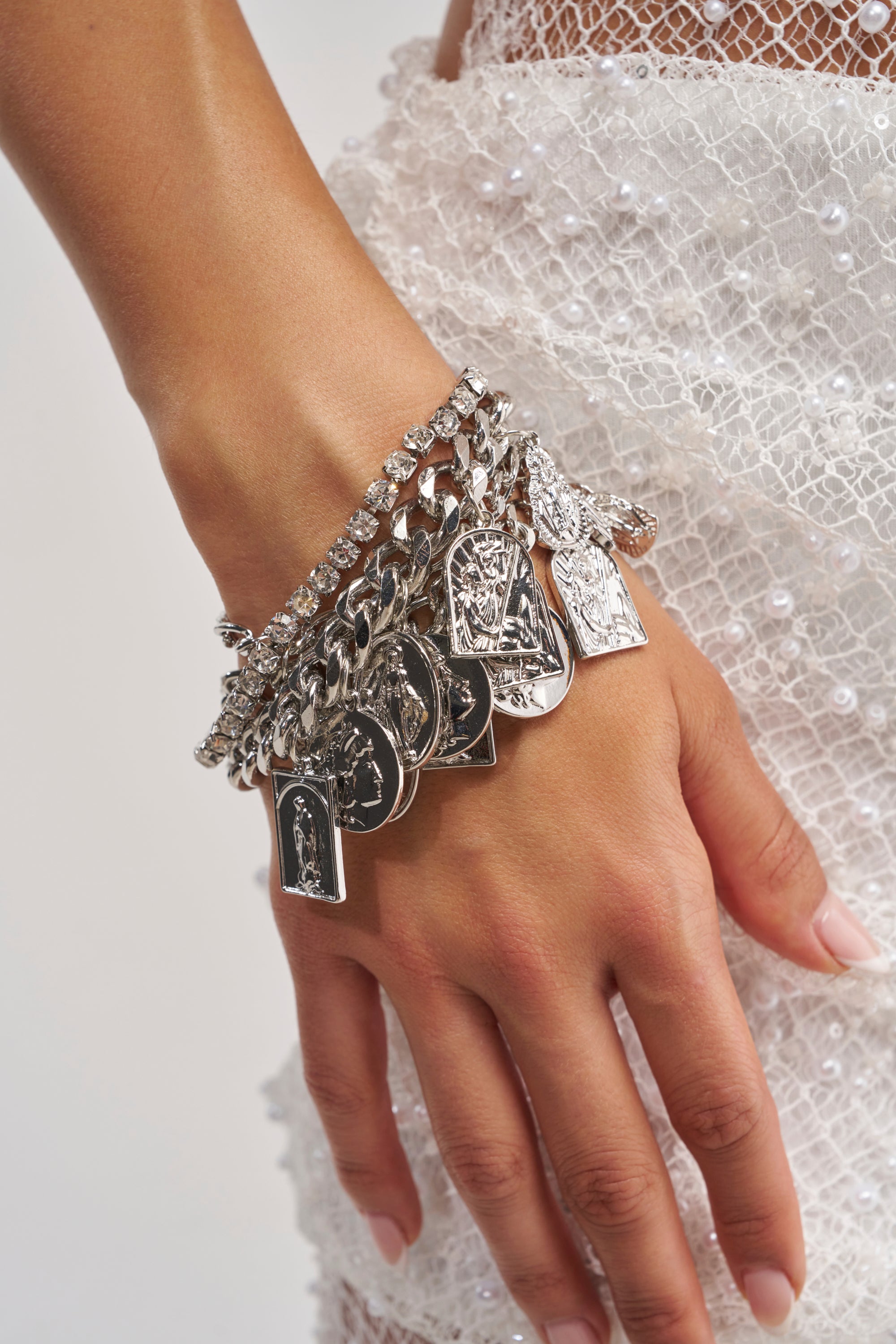 A hand adorned with the SAY YOUR PRAYERS BRACELET SET—layered silver chain bracelets featuring religious pendant charms—rests on a sheer, white, pearl-embellished fabric skirt, creating a striking display of festival jewelry that blends elegance with spiritual symbolism.