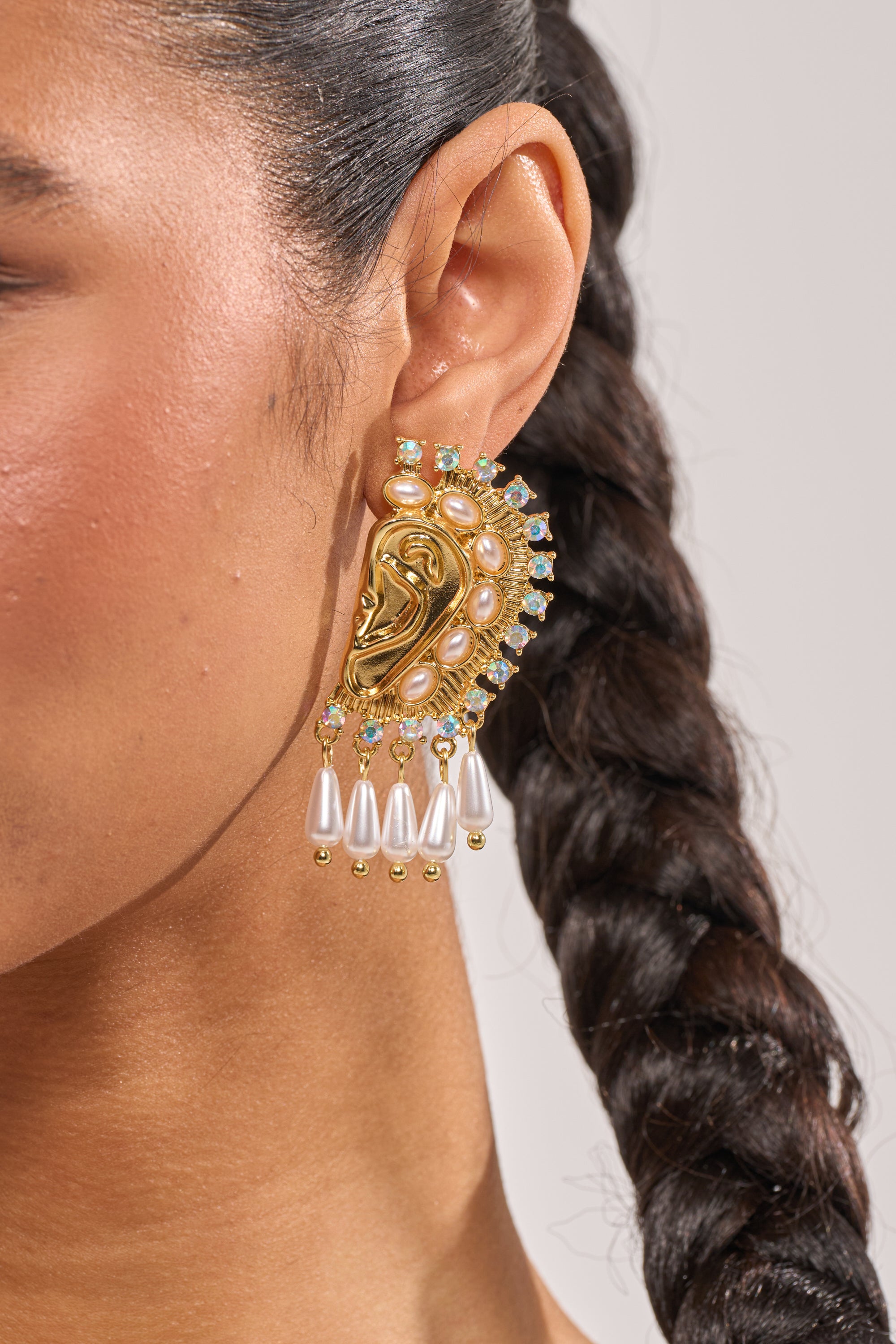 Close-up of a woman's ear wearing the I HEAR U EARRING—large, ornate statement earrings shaped like an ear with pearls, gemstones, and dangling pearl-like drops; her dark hair is braided. 1