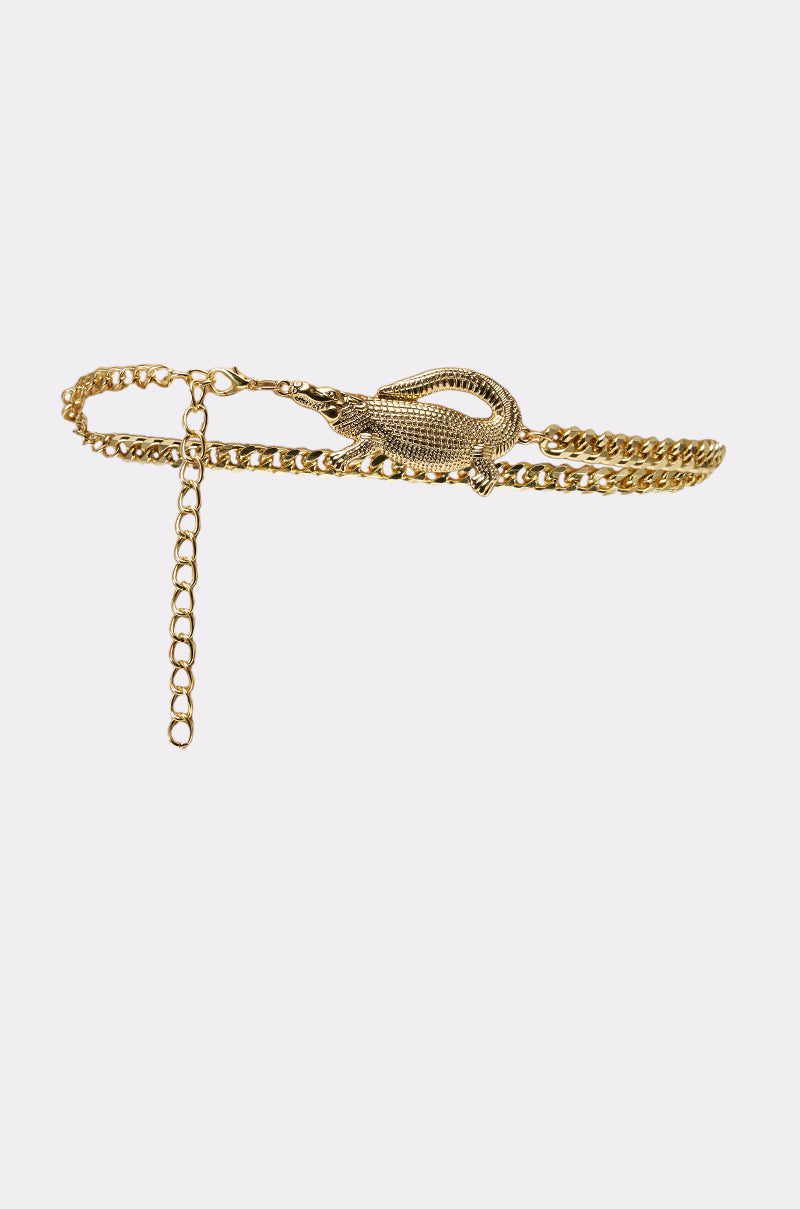 The BITE ME CHAIN BELT features a gold chain design with a bold crocodile buckle and an adjustable extender, creating a standout statement accessory. Shown against a plain white background. 1