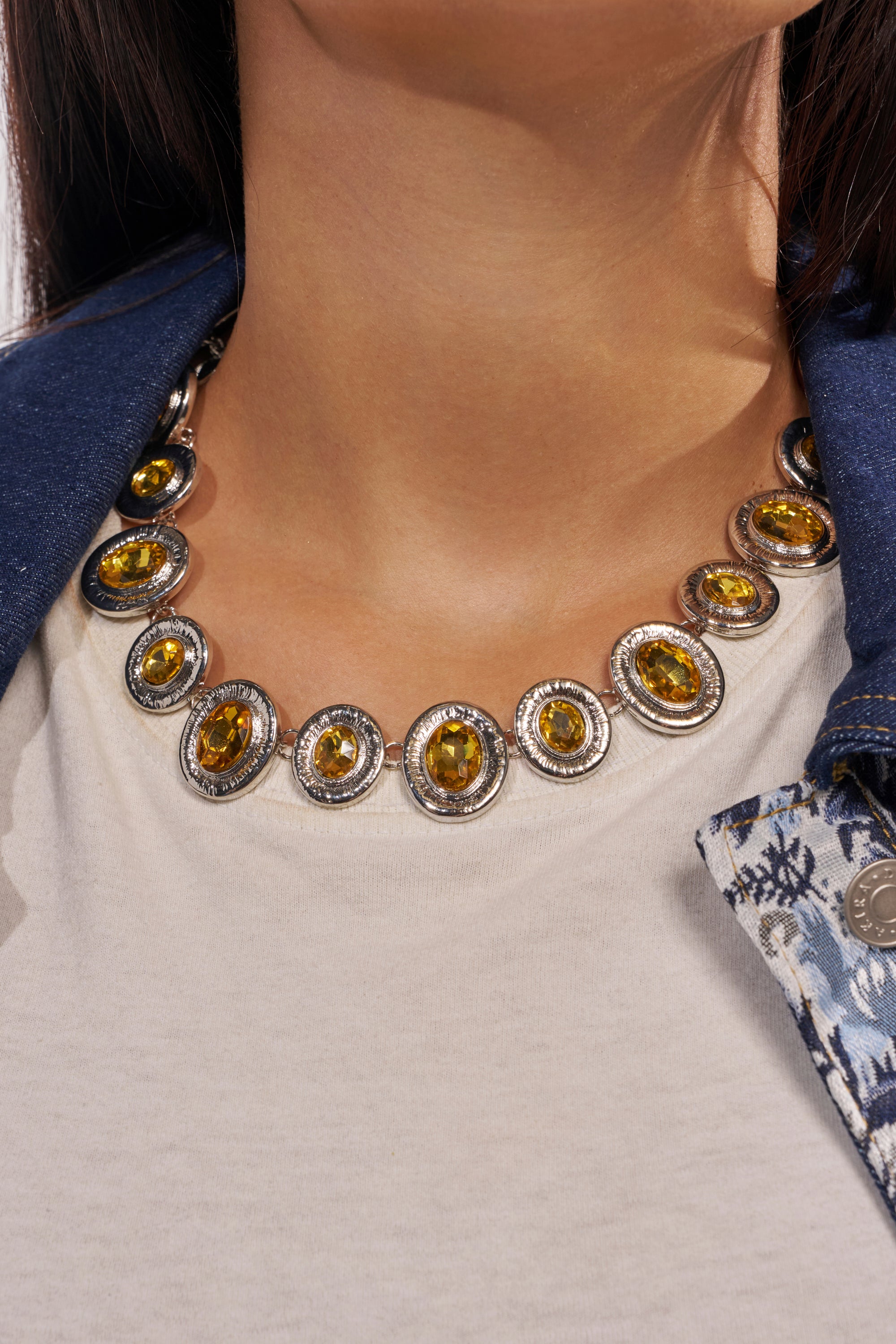 A close-up of a person wearing the FEEL IT NECKLACE, a silver-tone statement piece with yellow gemstones, styled with a white top and blue patterned jacket. 1