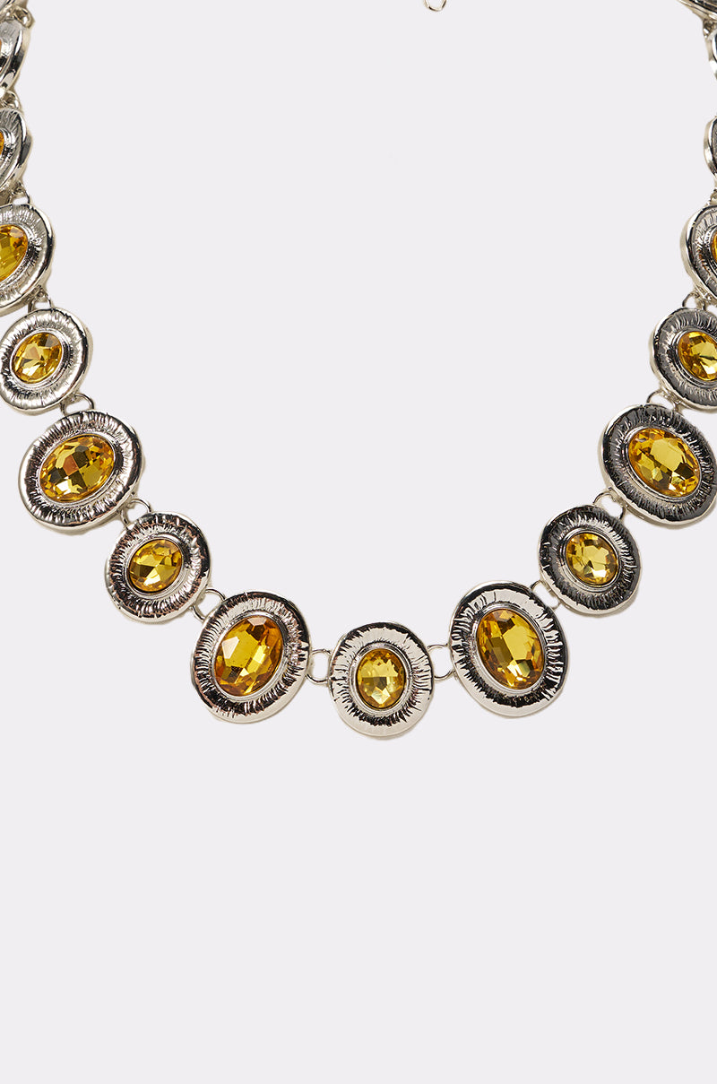 The FEEL IT NECKLACE is a silver-tone statement piece with oval and round yellow gemstones in ornate, symmetrical settings, shown close-up against a white background. 2