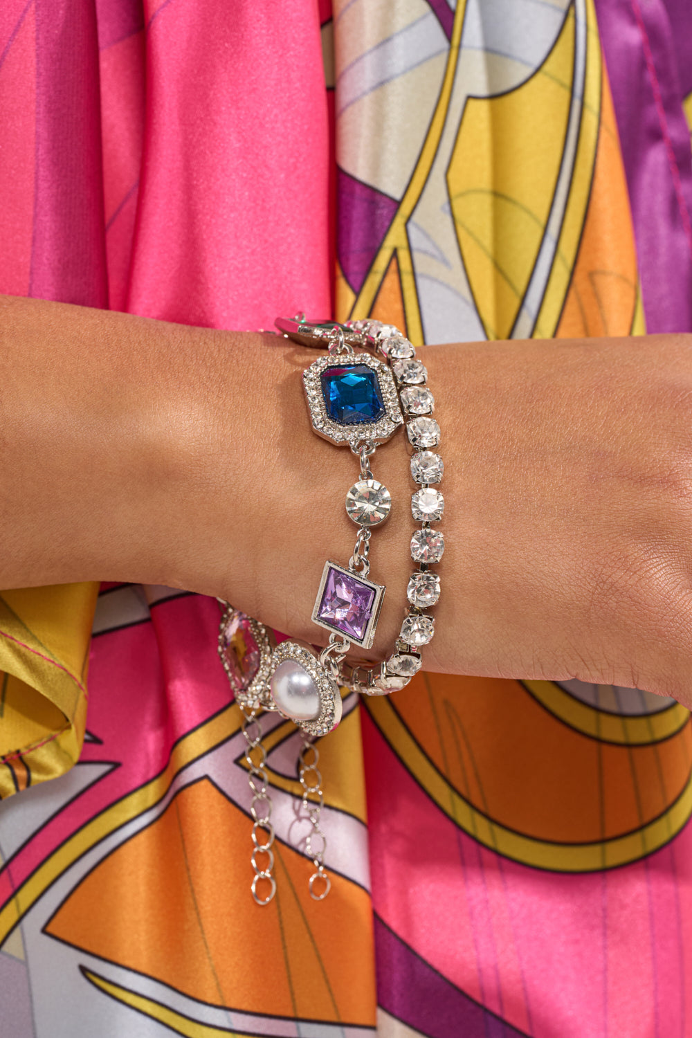 A close-up of a wrist adorned with the QUEEN APPROVED BRACELET SET, featuring a sparkling silver faux pearl and rhinestone bracelet with colorful gemstones and crystals, paired with a vibrant pink, yellow, and orange patterned outfit. 0