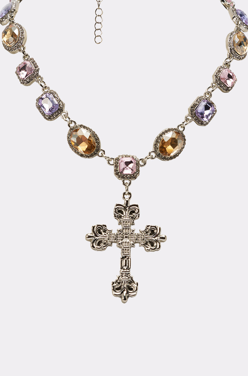 The MY LEGACY NECKLACE is a vintage-inspired statement piece with alternating gold, pink, and purple gemstones, featuring a large crystal cross pendant in the center. 3