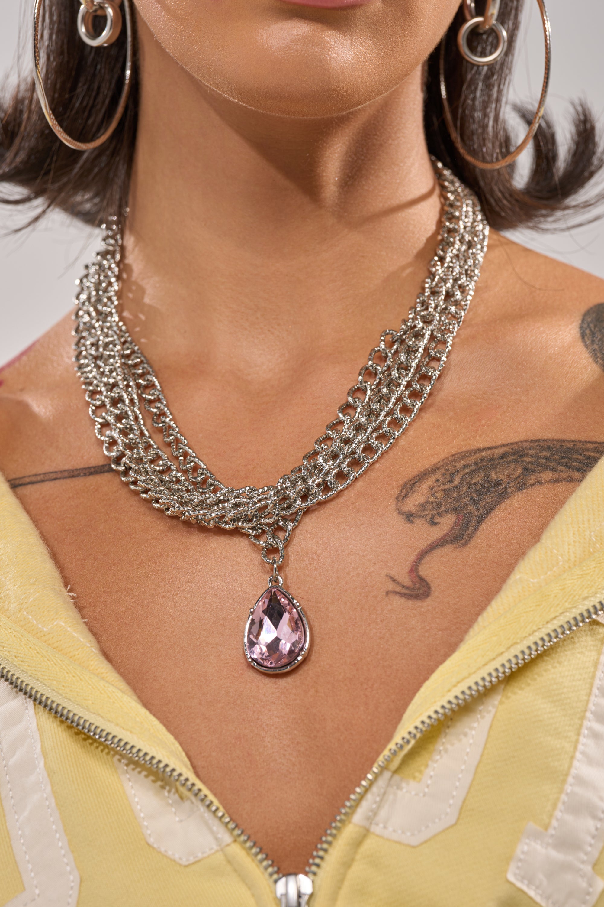 A person wears large hoop earrings, a yellow zip-up top, and the PRINCESS TREATMENT NECKLACE—a layered chain with a bold pink pendant. A snake tattoo is visible on their chest. 1