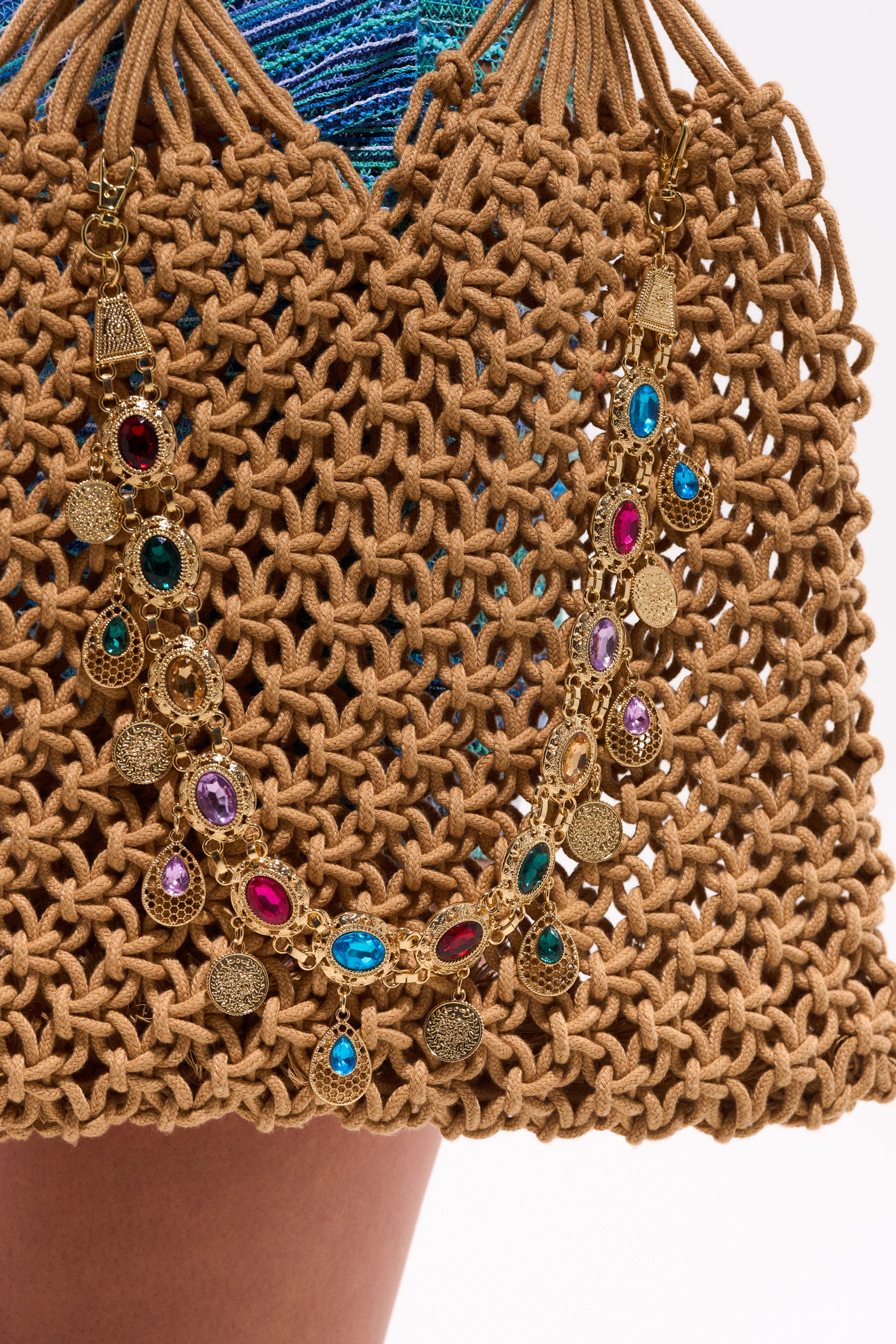 A close-up of a tan woven handbag adorned with the IM OUTSIDE PURSE CHAIN, featuring colorful gemstones and charms. Blue knitting appears partially in the background. 0
