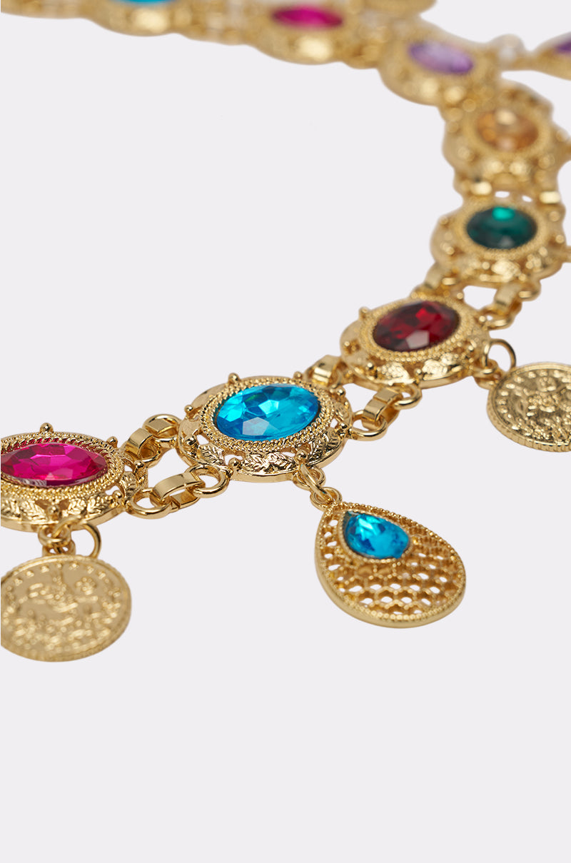 A close-up of the IM OUTSIDE PURSE CHAIN, a gold necklace with ornate circular and teardrop pendants featuring blue, red, and green gemstones, intricate detailing, and a polished finish—evoking an elegant jeweled purse chain. 2
