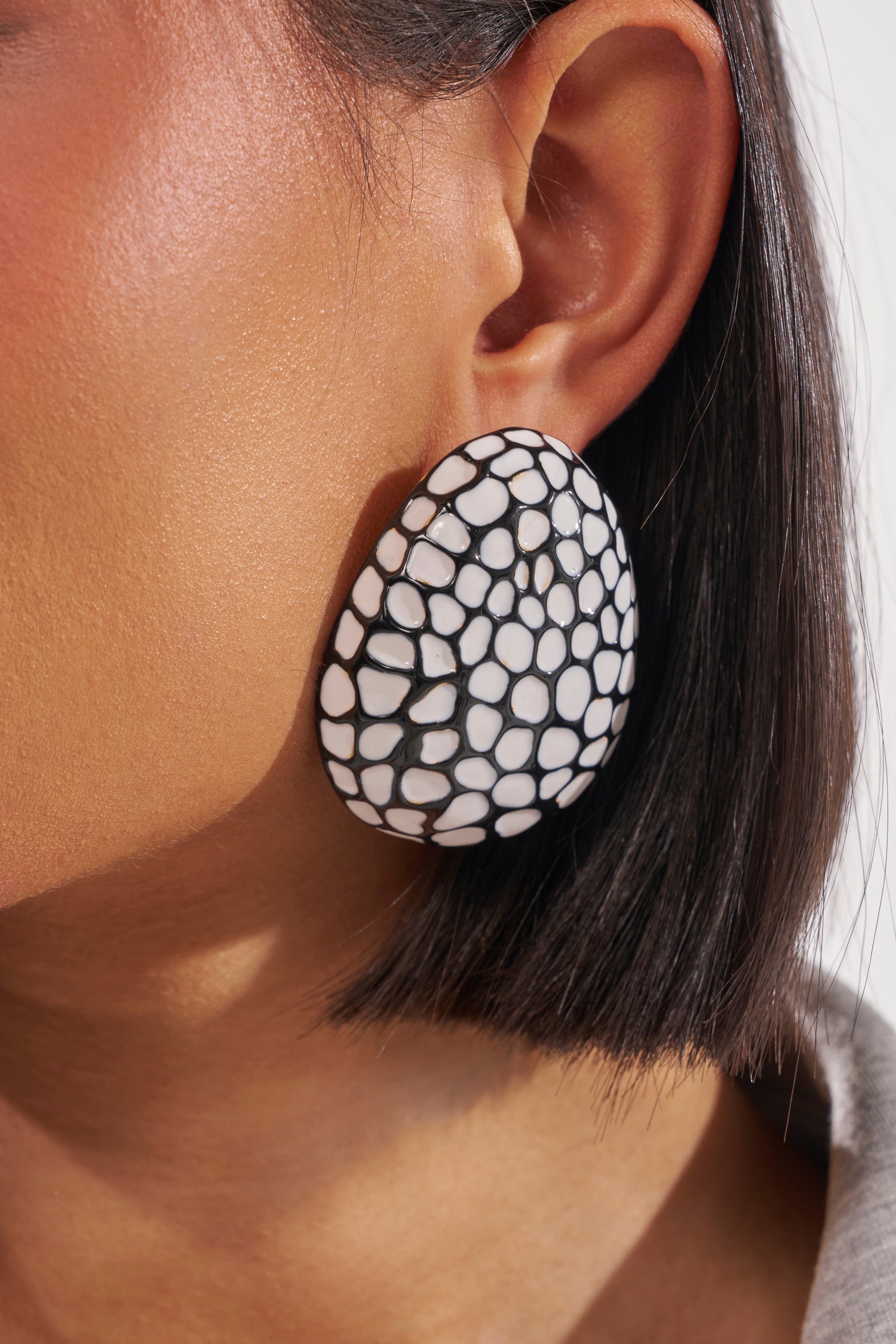 A close-up of a woman’s ear wearing the CANT LOSE EARRING—a bold, black-and-white mosaic stud with a cracked pattern—paired with straight, chin-length dark hair. 1