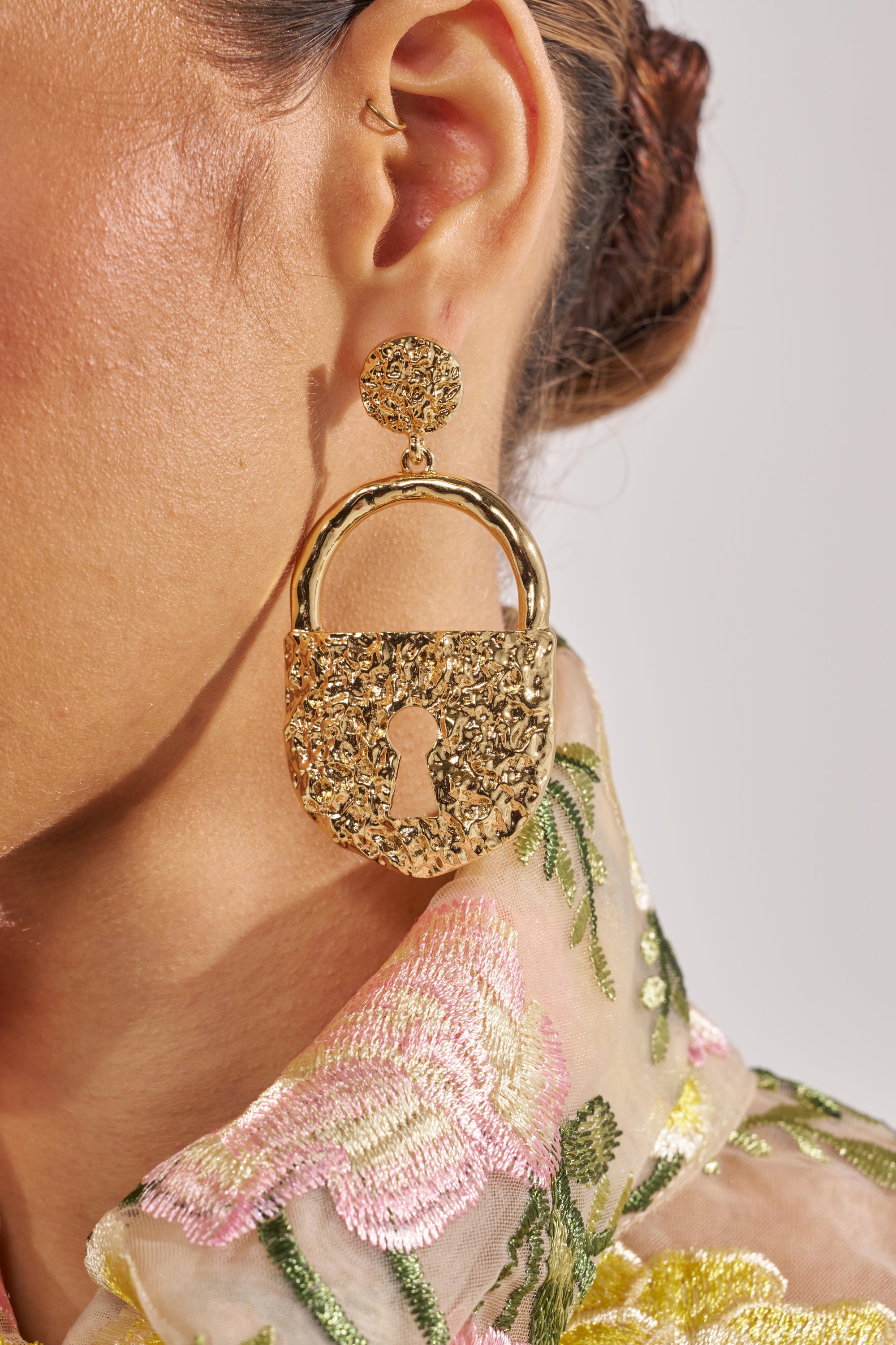 A close-up of a person’s ear wearing the ON LOCK EARRING—gold-tone, chunky drop earrings with a hammered texture and keyhole design—paired with sheer, floral embroidered fabric. 1