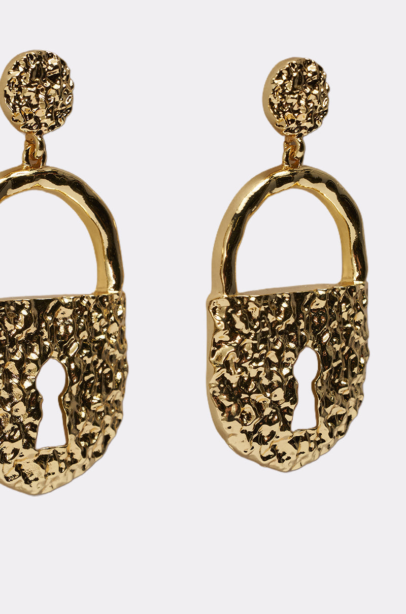 Close-up of the ON LOCK EARRING, featuring chunky padlock shapes with a hammered texture and keyhole cutout in gold-tone drops, set against a plain light background. 3
