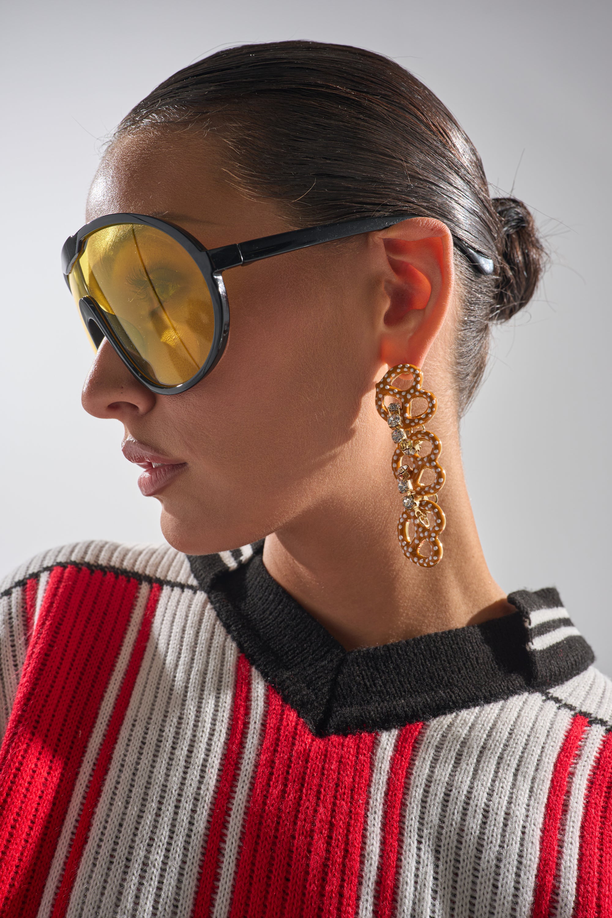 A woman with slicked-back hair wears large yellow sunglasses, the NEVER SALTY EARRING—a rhinestone chain style—and a red, white, and black striped sweater, photographed in profile against a plain background. 0