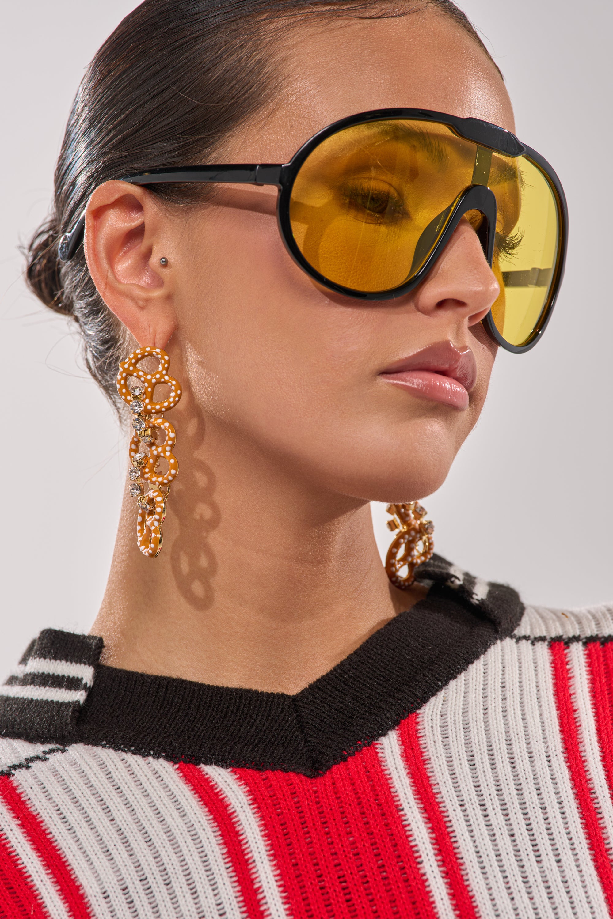 A woman with her hair pulled back and a calm expression looks to the side, wearing large yellow-tinted sunglasses, the NEVER SALTY EARRING, and a black, white, and red striped sweater. 4